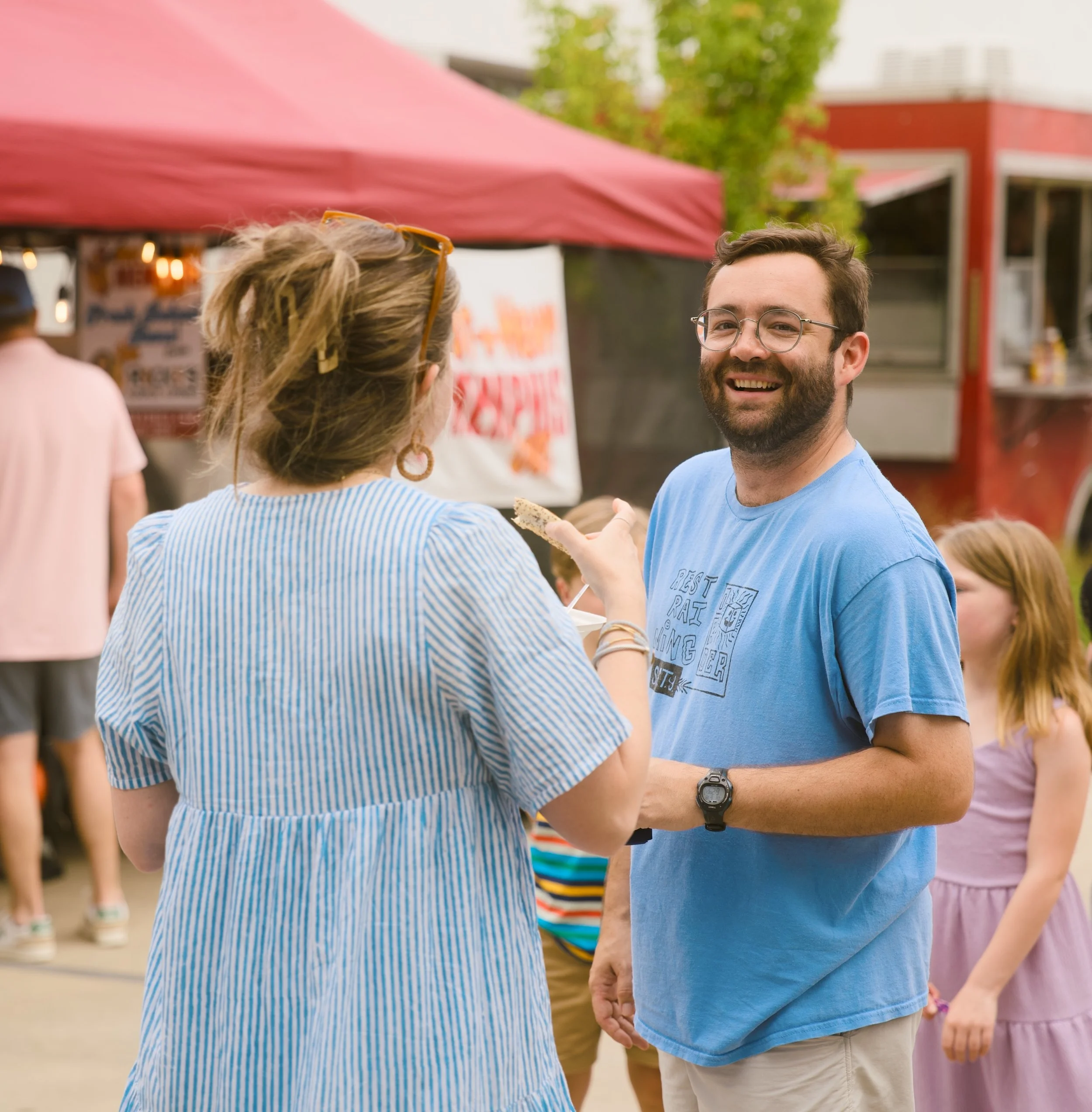 People at an outdoor event, focusing on a man with glasses smiling and talking to a woman with blonde hair wearing sunglasses on her head, in front of a red tent and greenery.