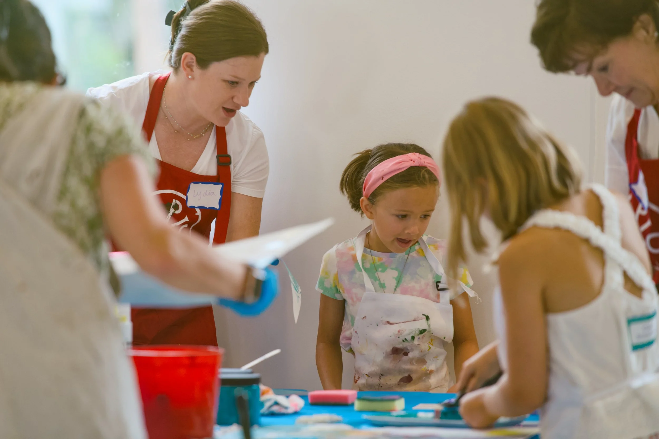 Children and adults participating in an arts and crafts activity in a brightly lit room, wearing aprons and focused on their projects.