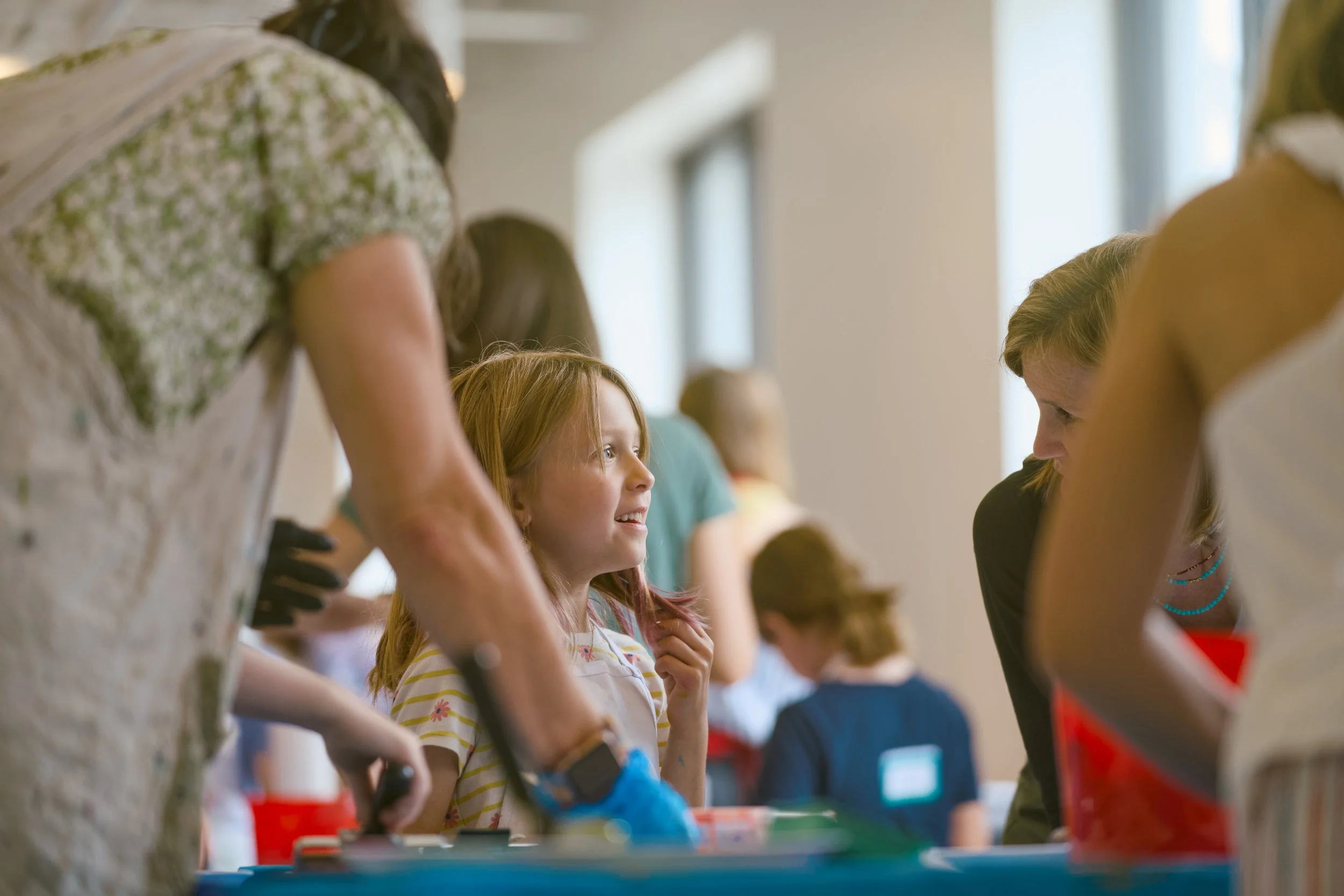 Children and adults in a line at a table, engaging in an educational activity or event inside a room with large windows.