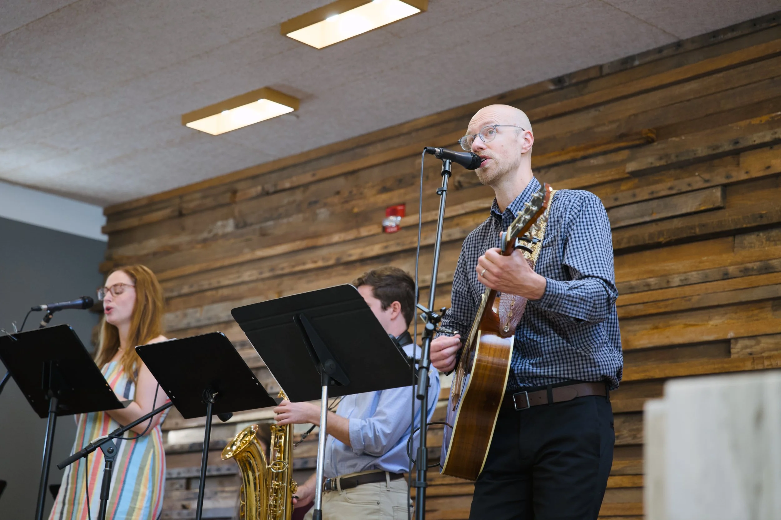 A group of musicians performing indoors, with a man holding an acoustic guitar and singing into a microphone. A woman with red hair and glasses is singing nearby, and a man with a saxophone is also visible. The background features a wooden wall and ceiling lights.