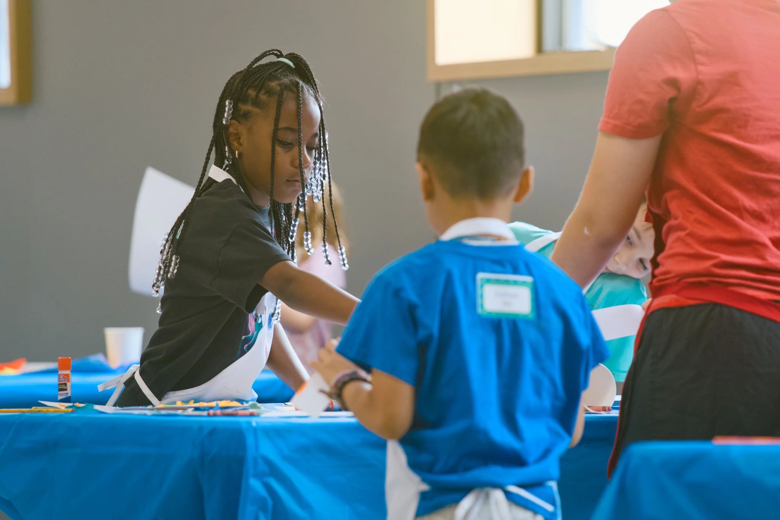 Children and an adult in a classroom setting, engaging in a craft activity at a table covered with a blue tablecloth, with windows letting in natural light.
