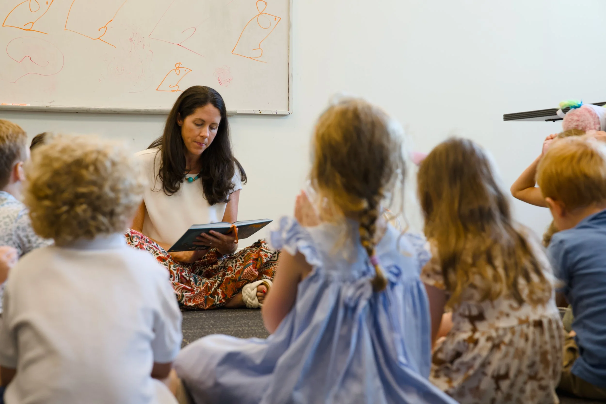 A woman sits on the floor reading a book to a group of children in a classroom, with a whiteboard behind her that has simple drawings of animals. The children are gathered around her, listening attentively.