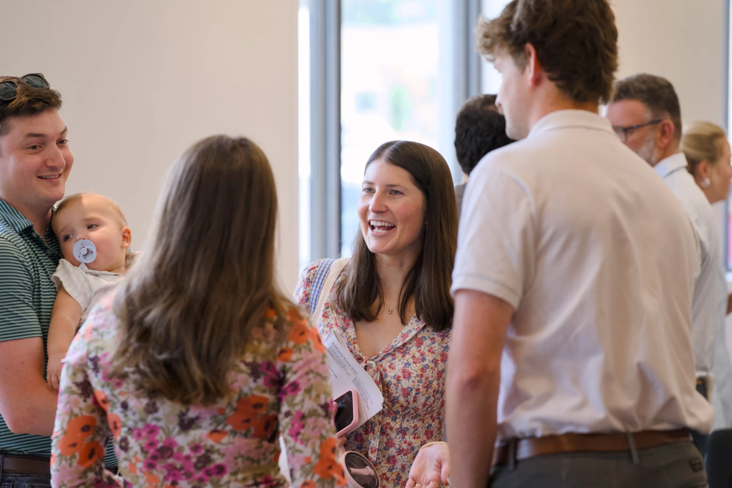 People engaging in conversation at an indoor social event, including men, women, and a young child with a pacifier, near a window.