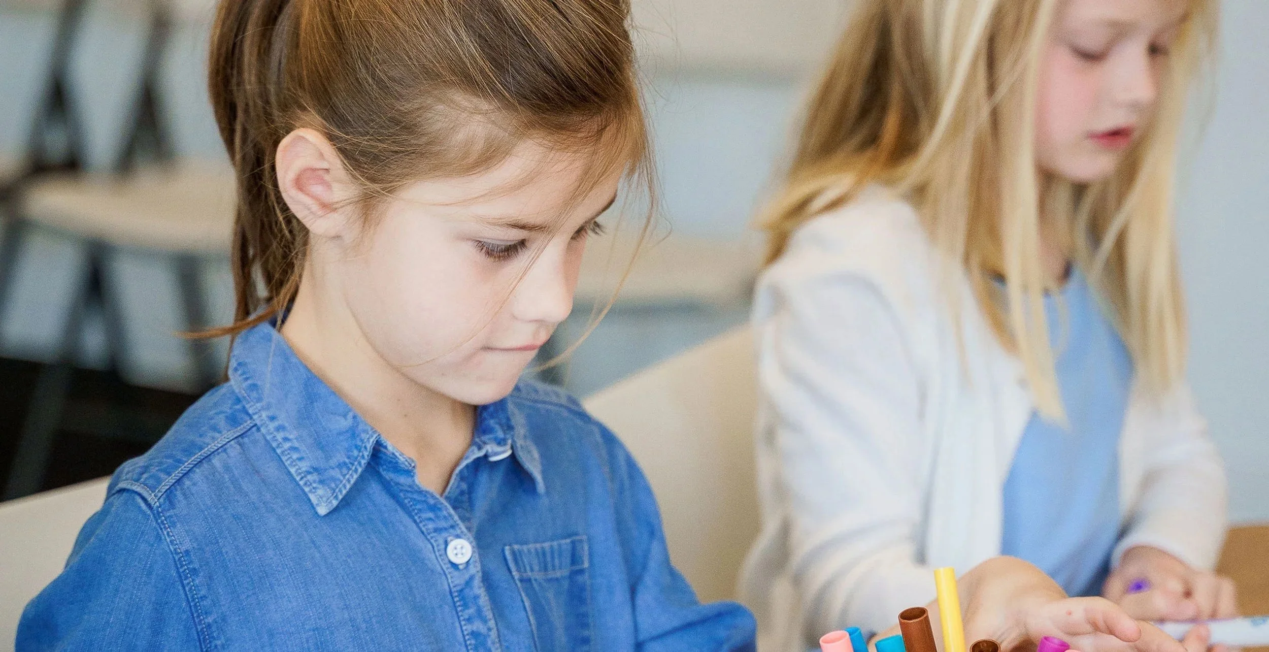 Two young girls sitting at a table, one with red hair and the other blonde, engaged in a craft activity with colorful markers.