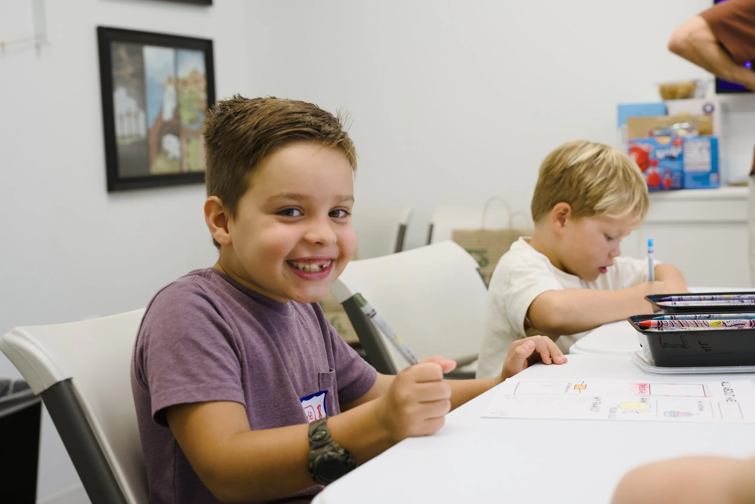 Young boy smiling while sitting at a table, working on a coloring project with markers and papers, in a classroom setting.
