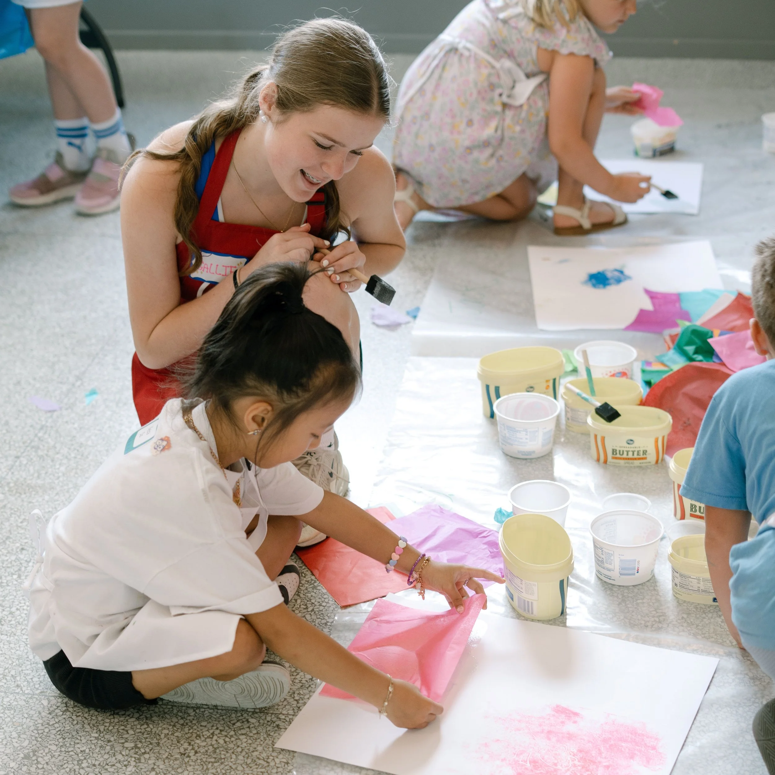 Young girl in a white uniform and colorful bracelets drawing with pink and blue paint on large sheets of paper on the floor, while adults and children work on similar creative projects nearby.