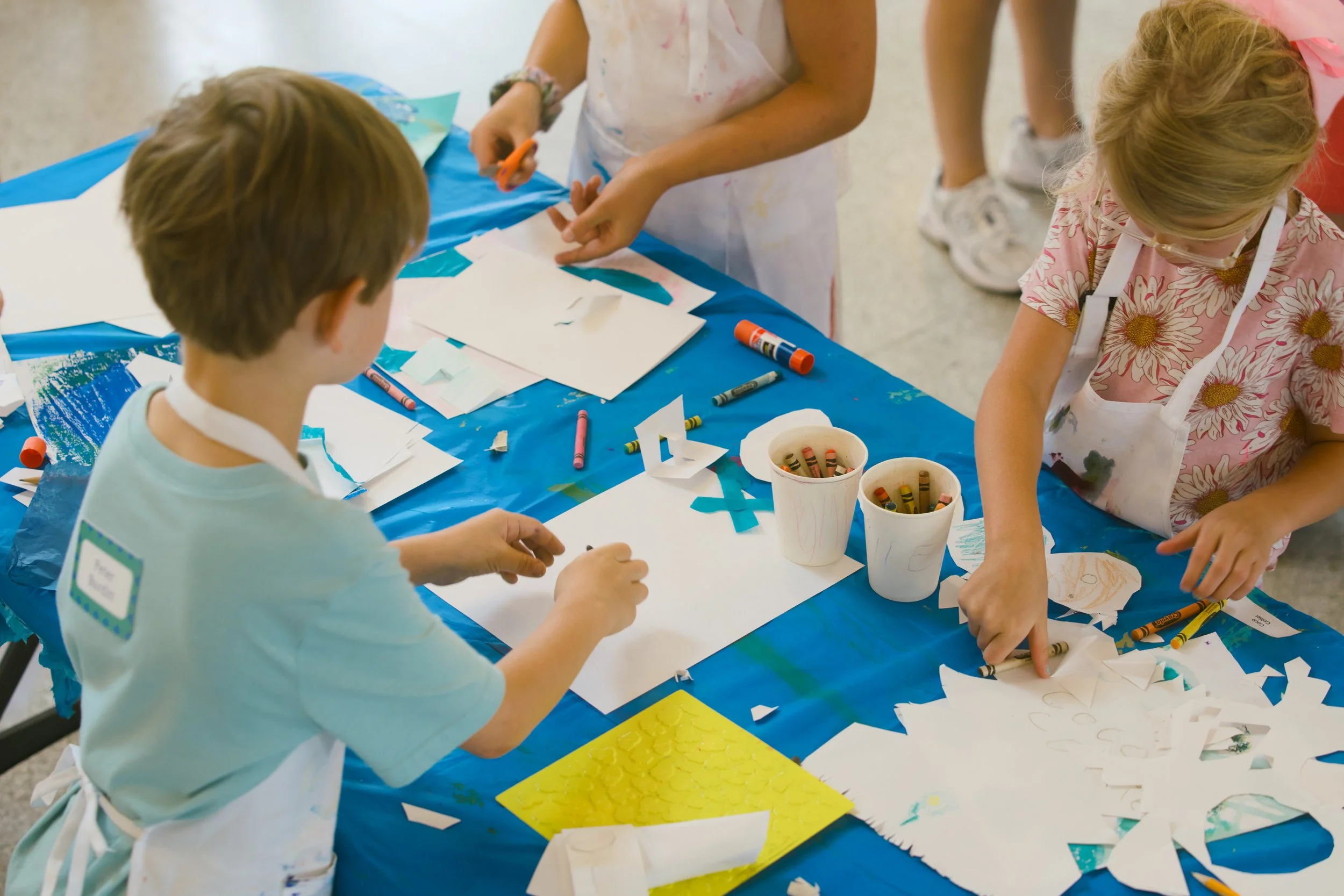 Children engaged in arts and crafts activity around a table covered with blue tablecloth, using scissors, crayons, and paper.