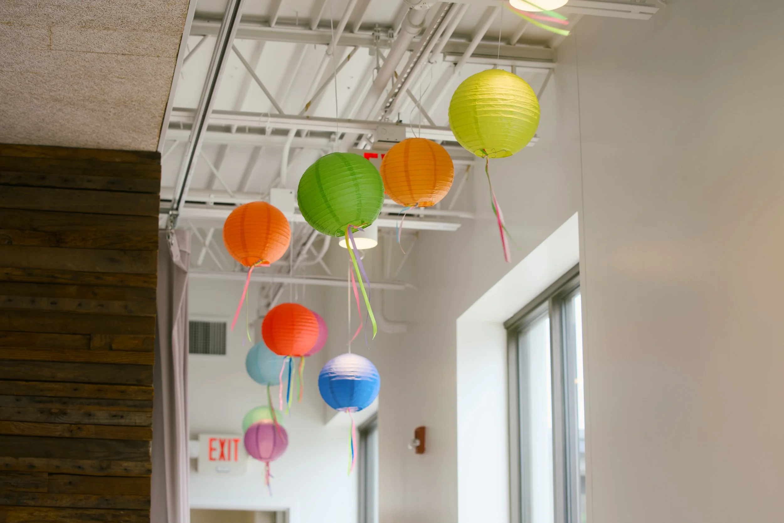 Colorful paper lanterns hanging from the ceiling in an indoor space with large windows and exposed ceiling beams.
