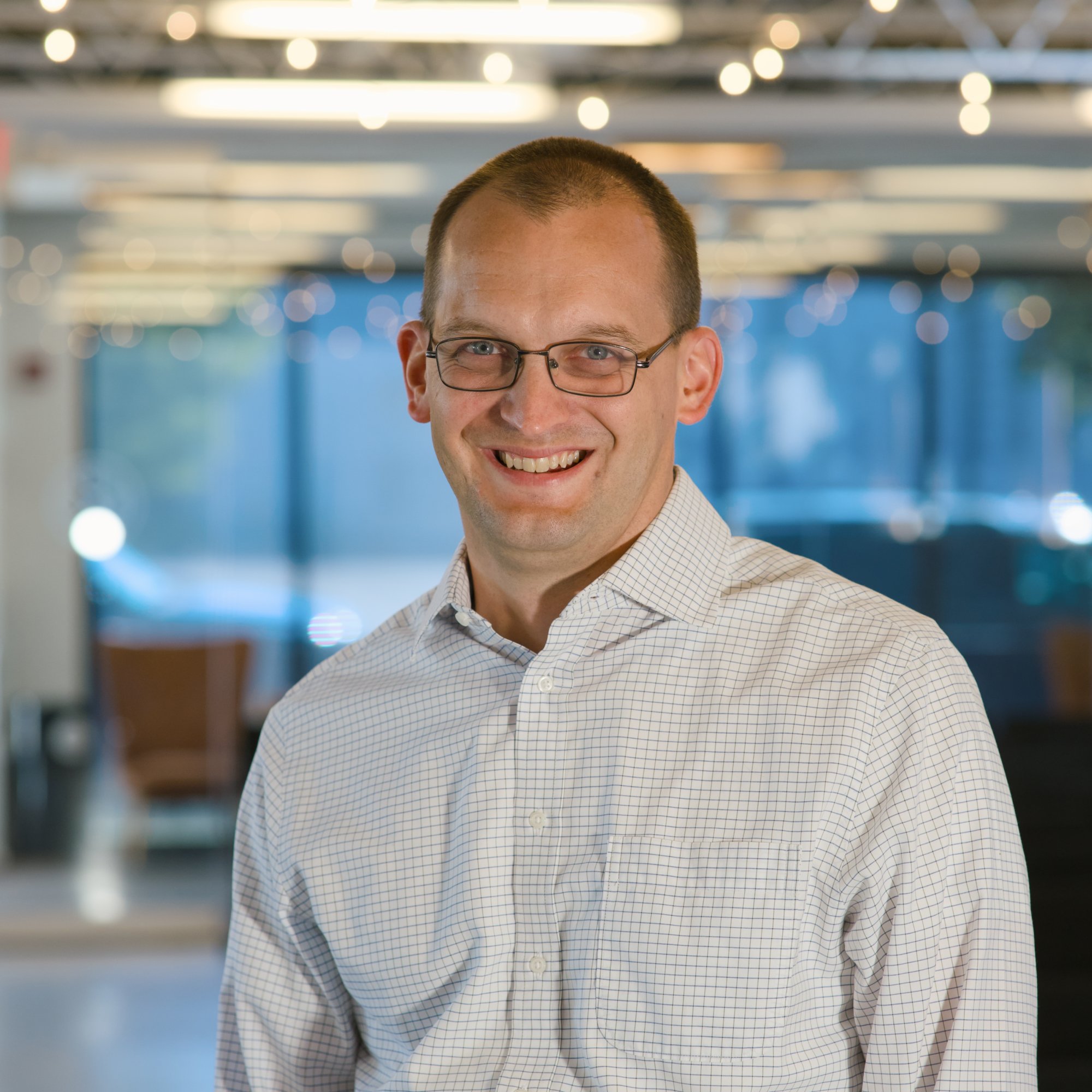 A man with short hair, glasses, and a checkered shirt smiling in an indoor setting with blurred lights in the background.