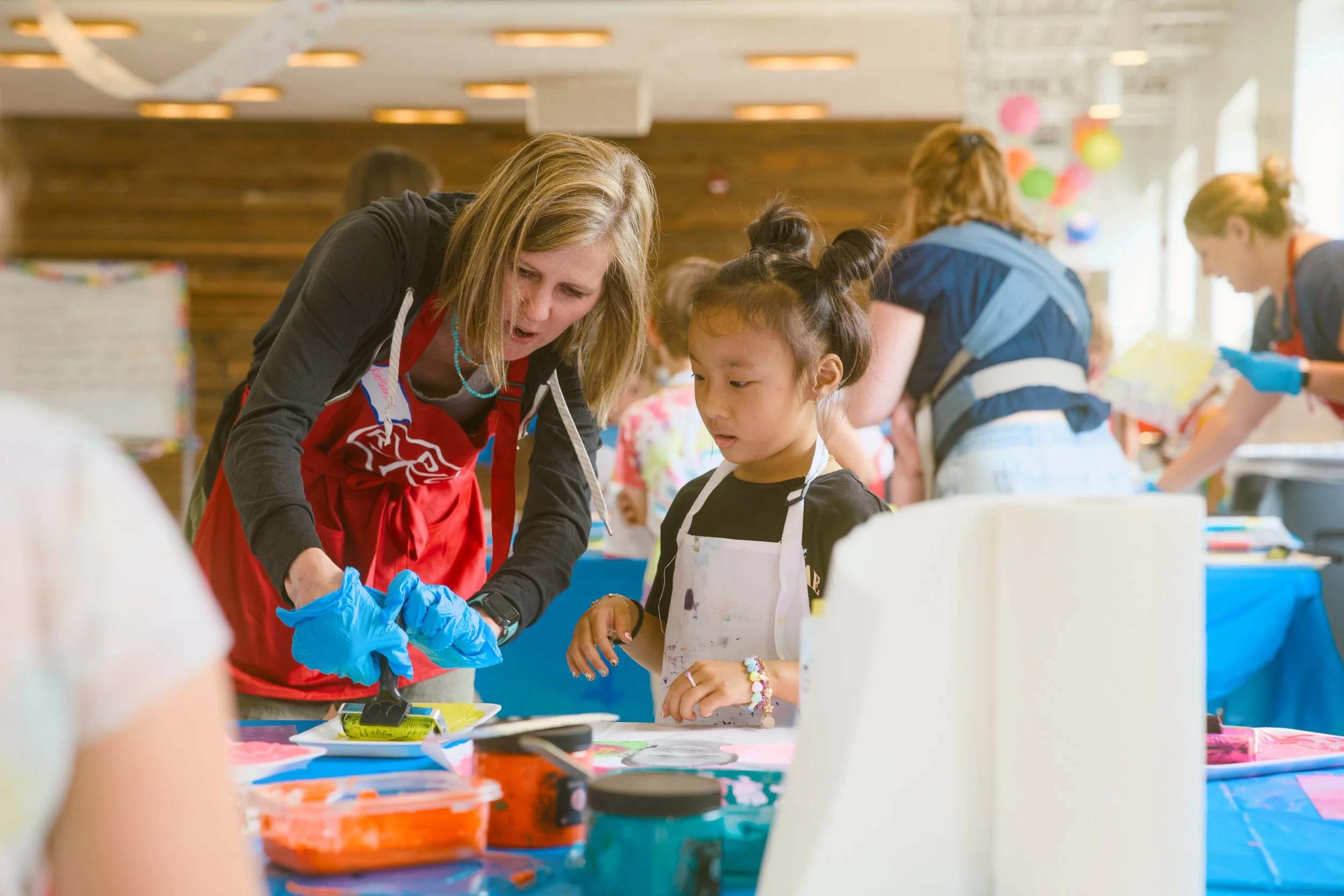A woman in a red apron and blue gloves helps a young girl with dark hair styled in buns, both working on an arts and crafts project at a table. Several people are in the background in a well-lit, decorated room.