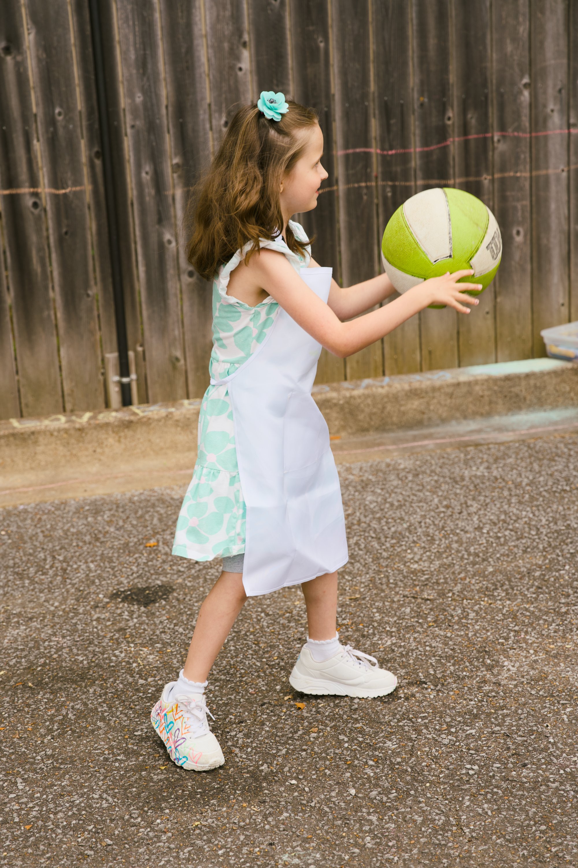 A young girl with brown hair wearing a blue flower hair accessory, a patterned sleeveless dress, an apron, and white sneakers with colorful heart designs. She is holding a green and white volleyball, standing outdoors on a gravel surface in front of 
