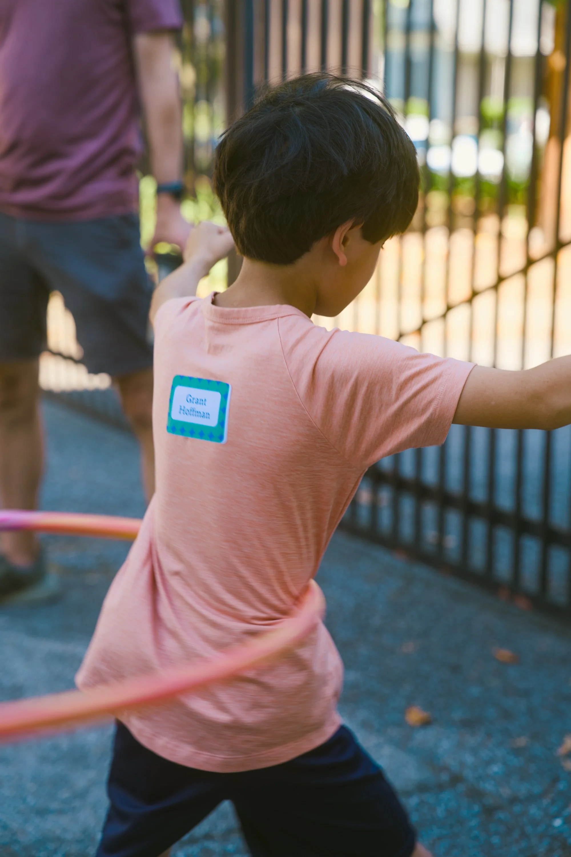 A young boy wearing a pink T-shirt with a name tag that says 'Grant Hoffman' is participating in a jump rope activity outdoors near a black fence, with another person partially visible in the background.