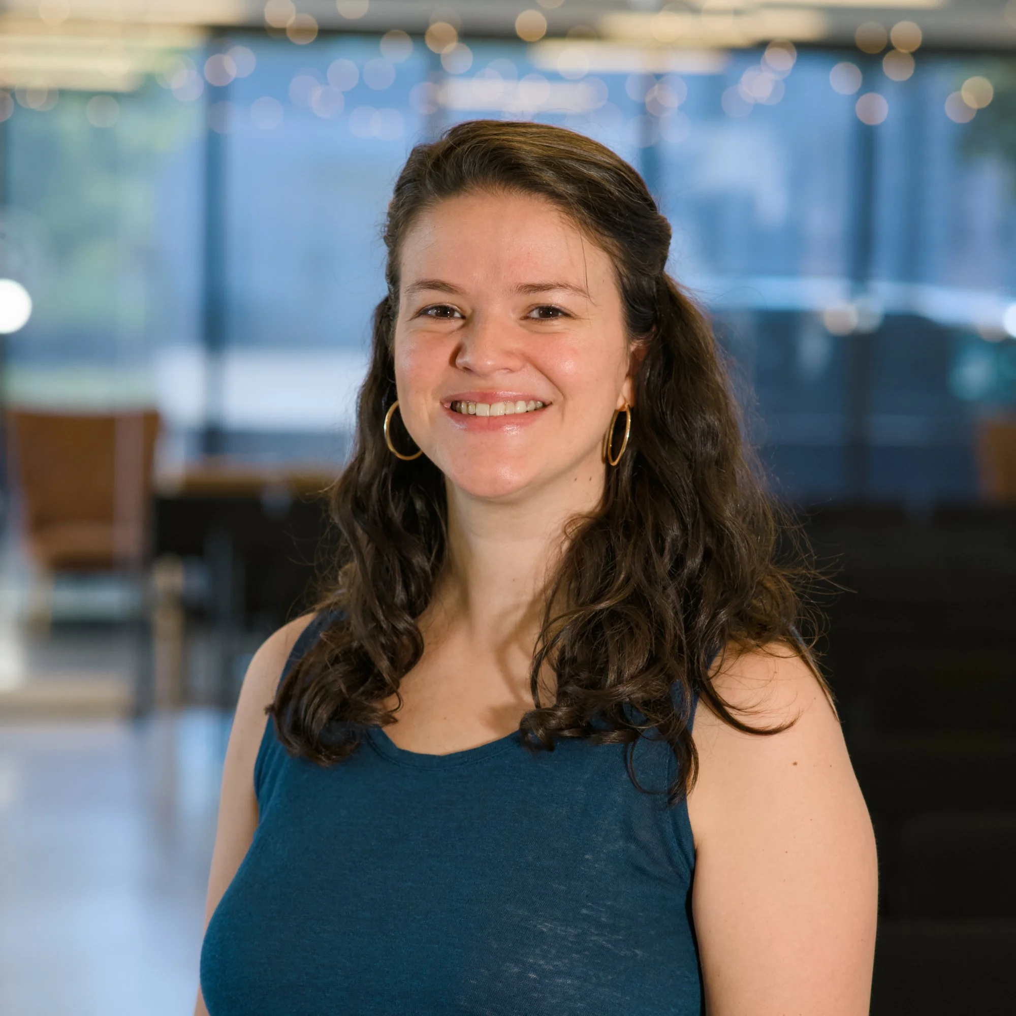 A smiling woman with wavy brown hair wearing large gold hoop earrings and a dark blue sleeveless dress, standing in front of an out-of-focus background with blue and bokeh lights.