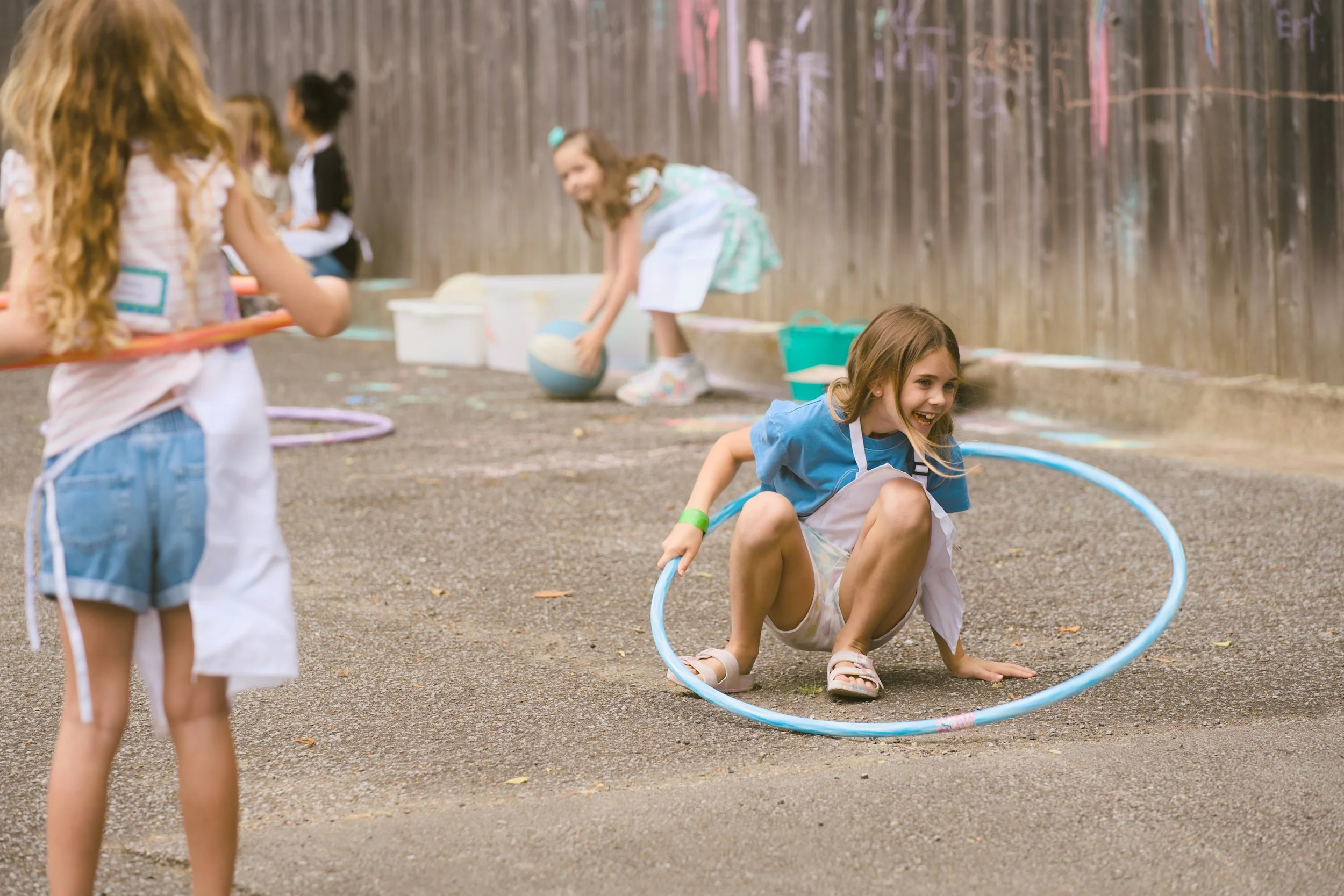 Children playing outdoors, one girl crouching with a hula hoop around her, others playing with a basketball and hula hoops, on a gravel surface with a wooden fence in the background.