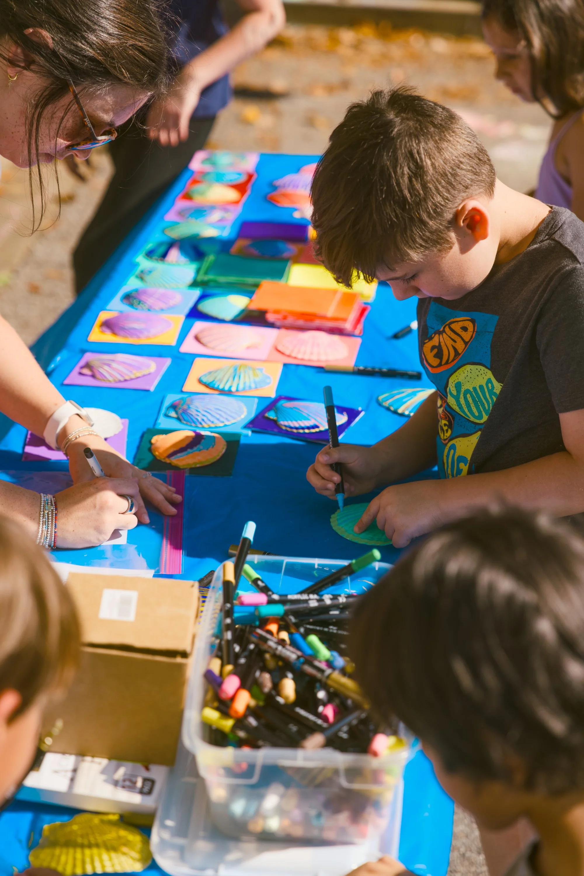 Children and adults gathered around a table outdoors, coloring seashells with markers for an arts and crafts activity.