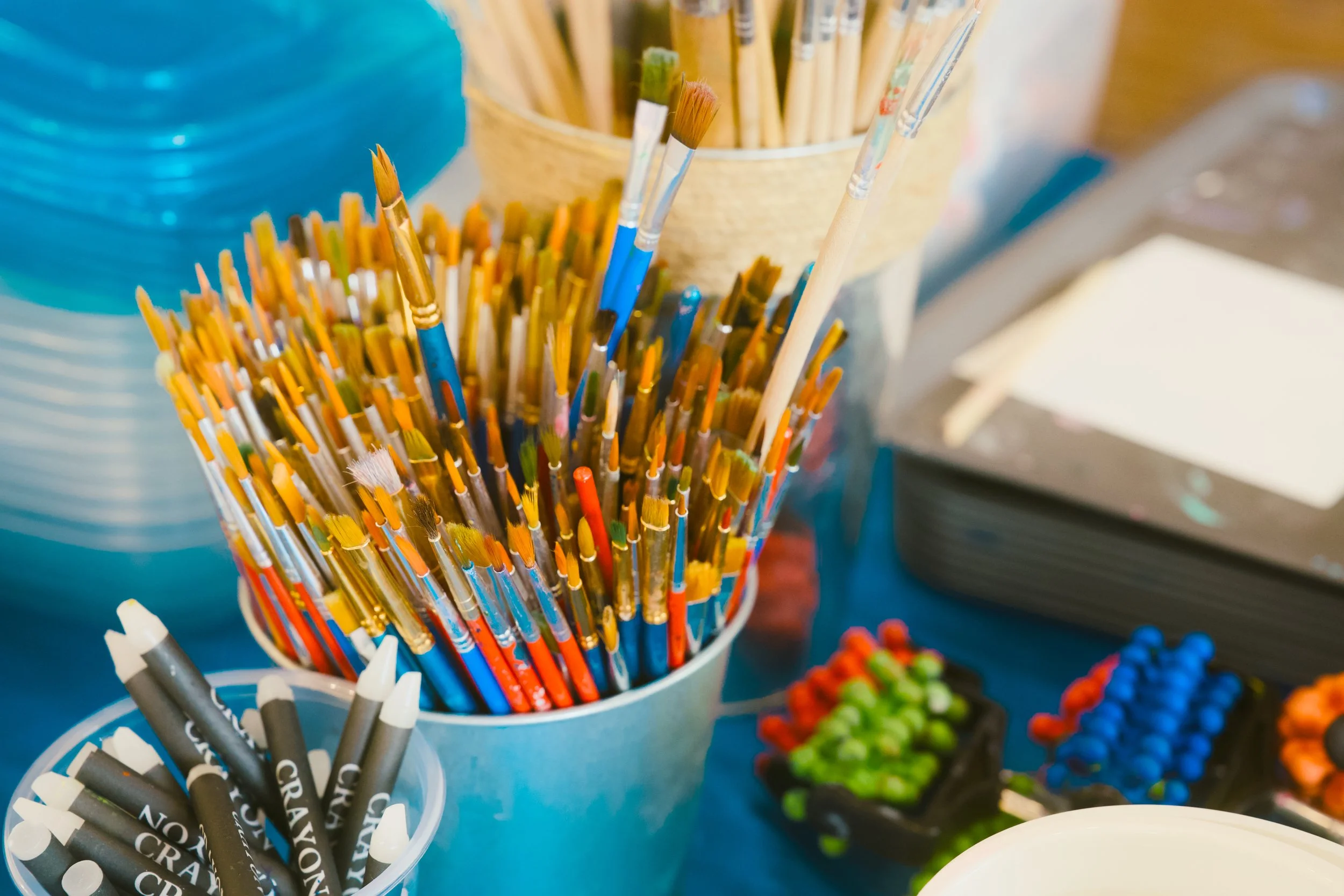 Various paintbrushes and markers in containers on a table, with some colorful objects in the background.