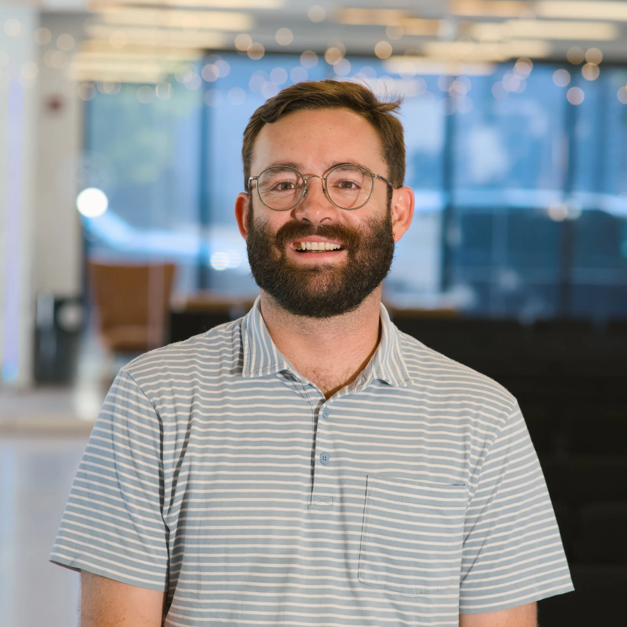 Close-up of a man with glasses and a beard, smiling, wearing a striped collared shirt, in an indoor setting with blurred golden lights and windows in the background.