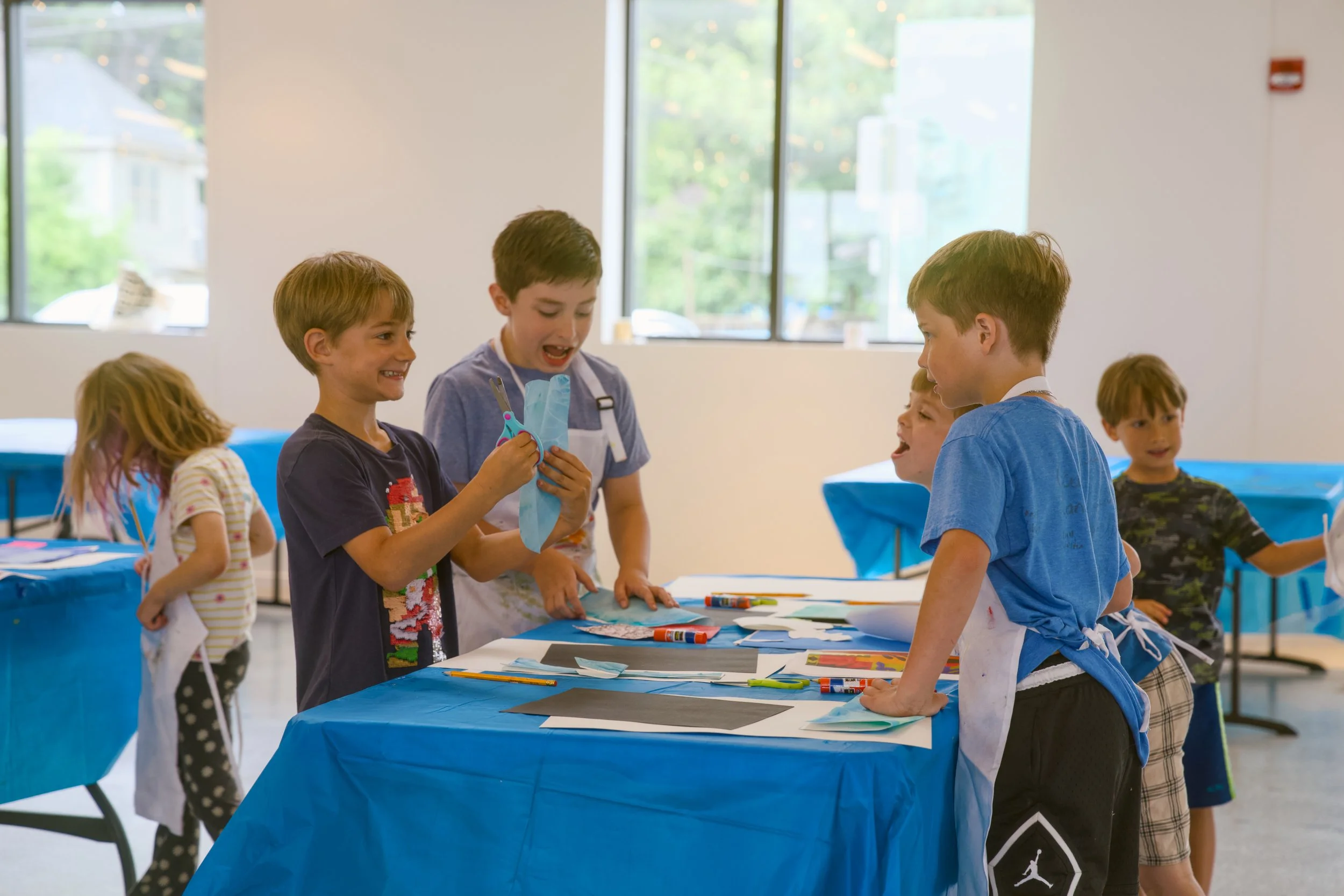Children gathered around a table at an art activity, with some holding art supplies, in a bright room with large windows.