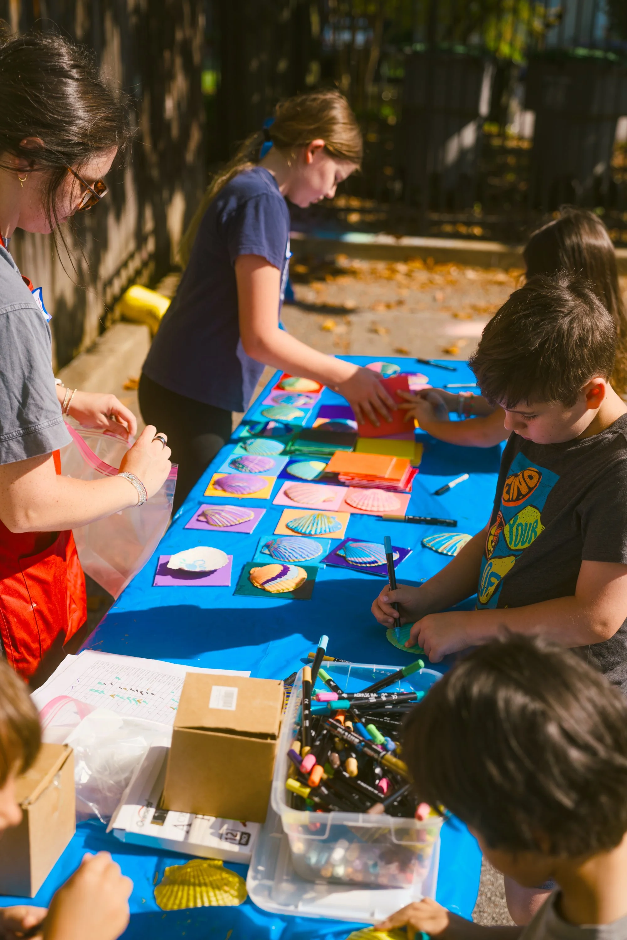 Children and adults gathered around a table outdoors, engaging in arts and crafts activities with colorful paper, markers, and shells.