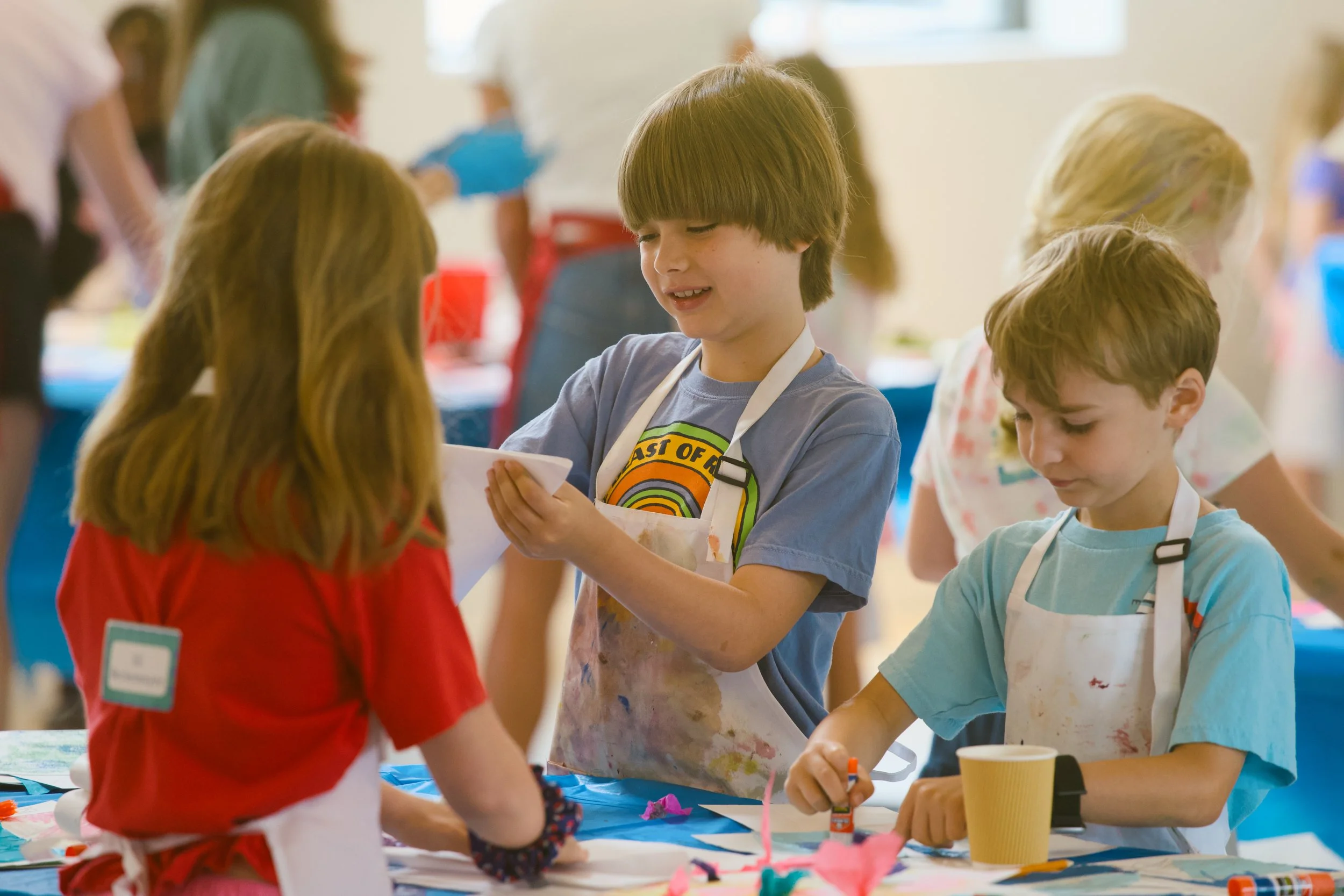 Children wearing aprons and engaging in arts and crafts activities at a table, with art supplies and paper in a bright, lively classroom setting.