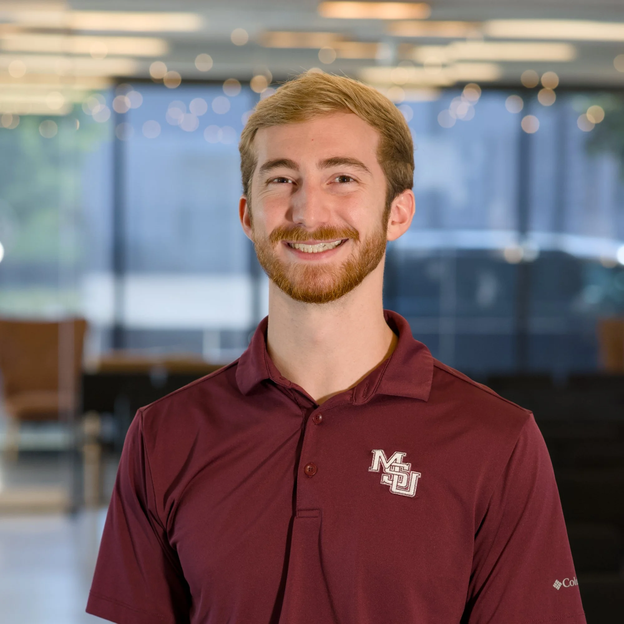 Young man with red hair and beard smiling, wearing a maroon collared shirt with a logo on the chest, in an indoor setting with blurred background lights.