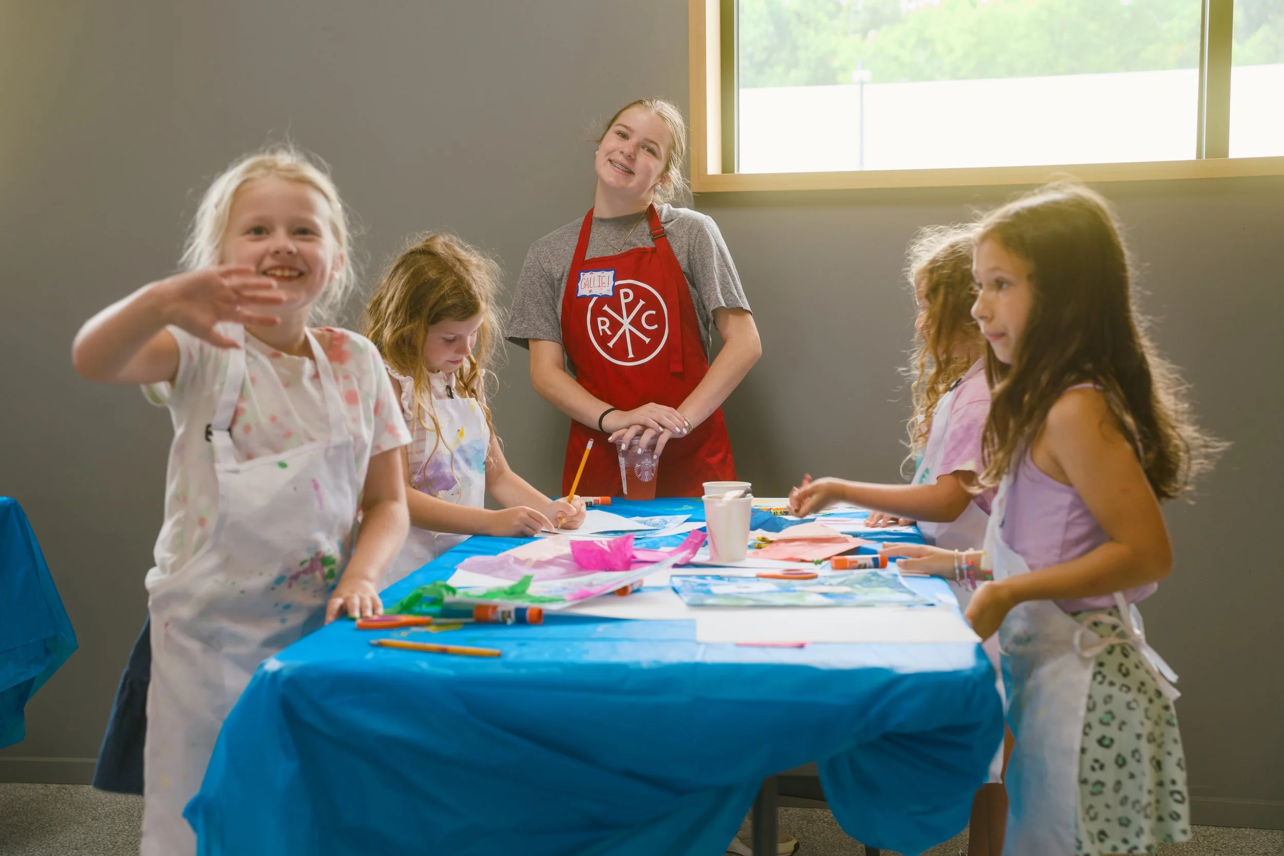 A group of young girls and a teenage girl, smiling and engaging in arts and crafts activities at a table covered with a blue tablecloth, in a bright room near a window, with some girls wearing aprons.