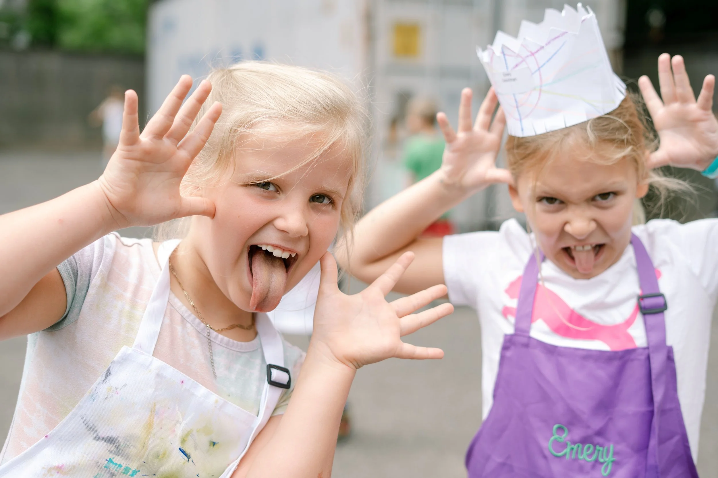 Two young girls at an outdoor event making funny faces and posing with their hands up, one wearing a crumpled paper crown.
