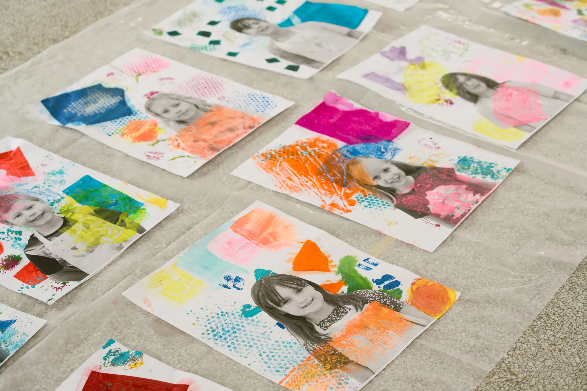 Multiple black-and-white portraits of children printed on paper, overlaid with colorful painted patterns and designs, laid on a tiled floor.