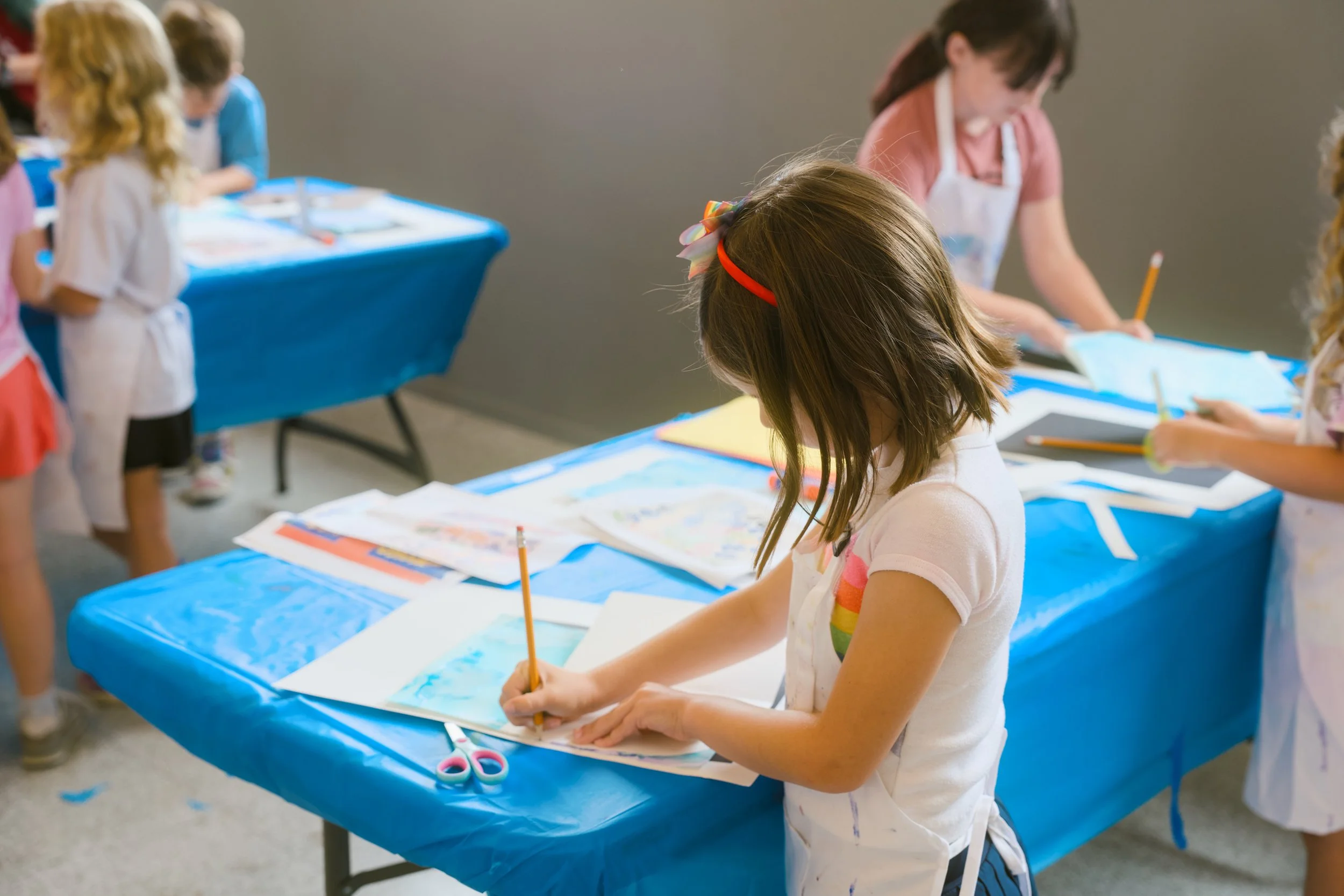Children, including a girl with a rainbow-striped bow in her hair, participating in a crafting activity at a table covered with a blue tablecloth, during an indoor art event.