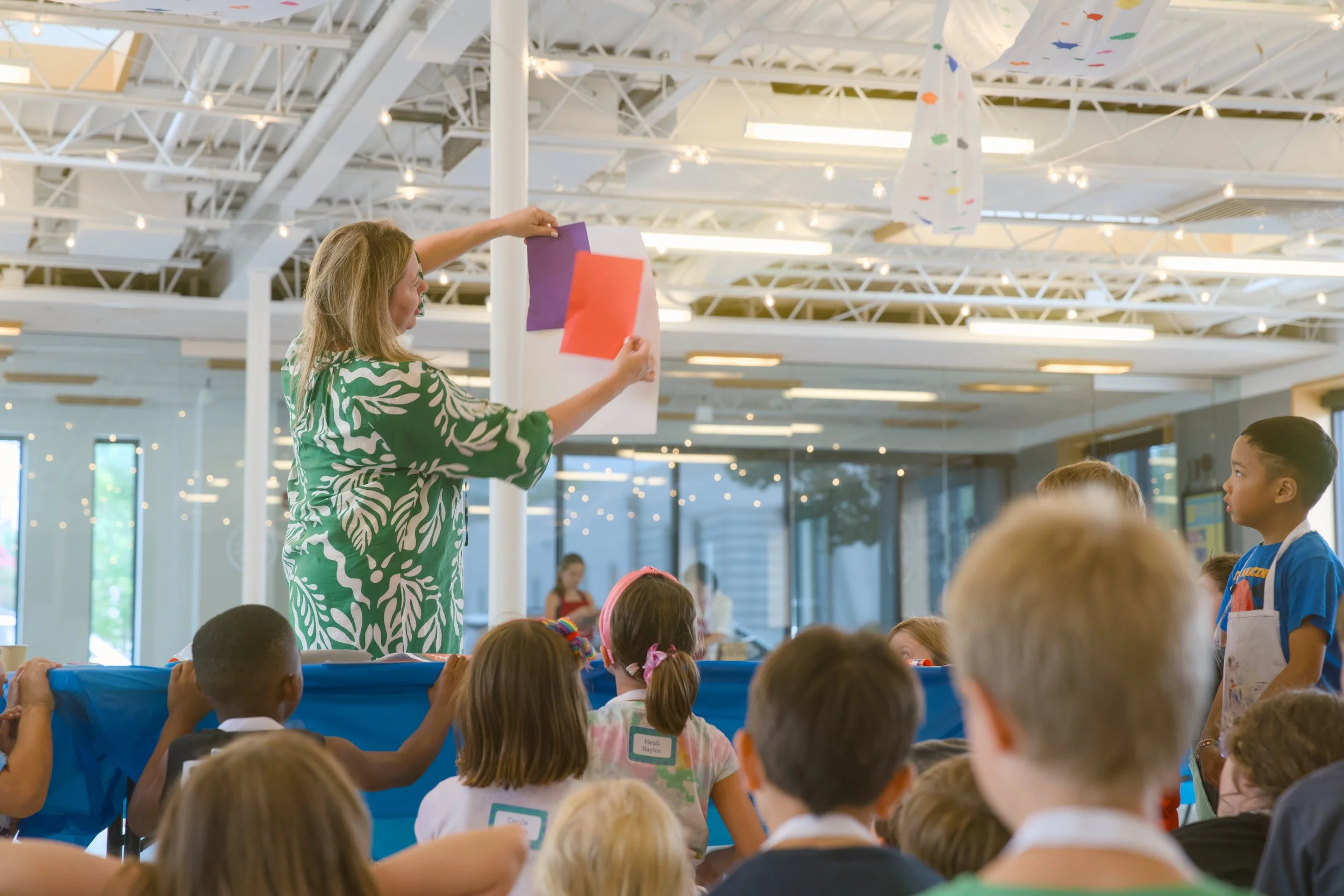 A woman in a green, patterned shirt is placing colorful paper on a pole in front of a group of children inside a bright, sunlit room with high ceilings and string lights. The children are standing and appear attentive, some holding a blue banner.