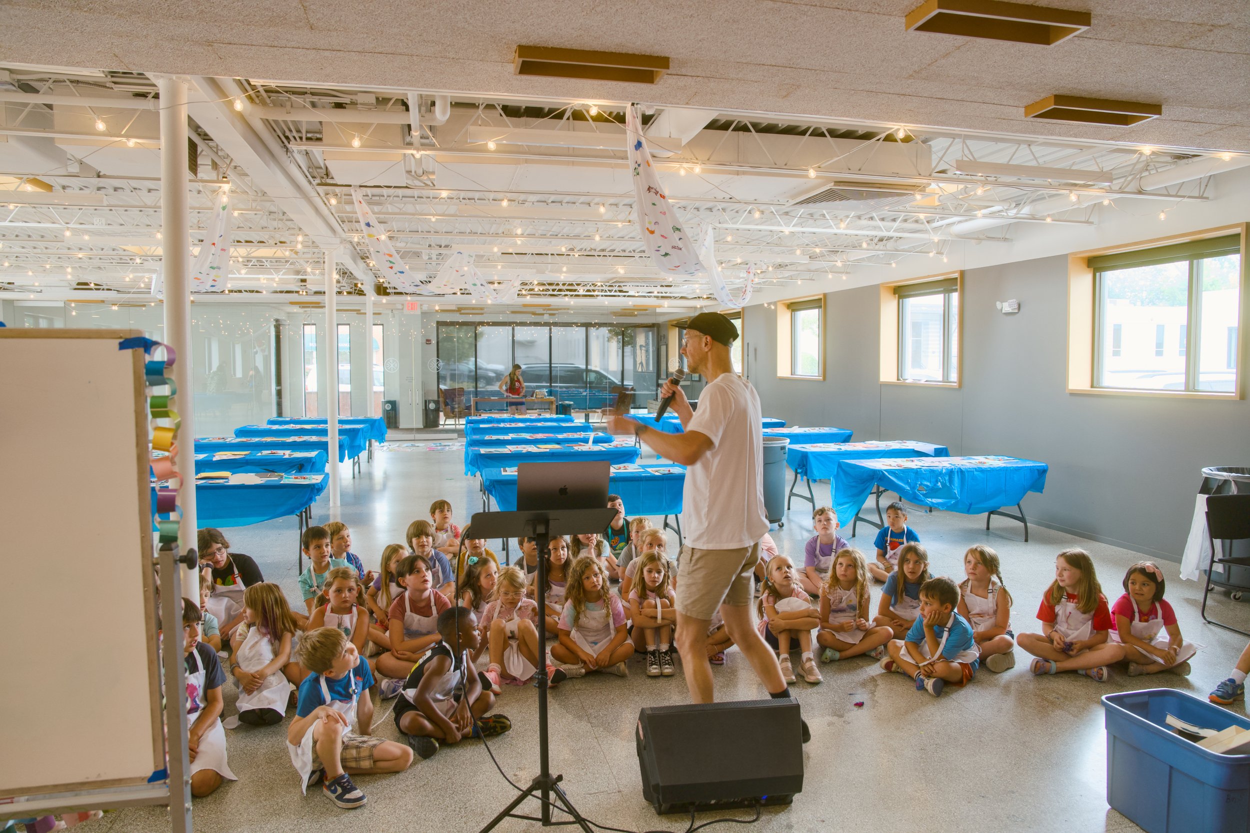A man with glasses and a cap stands in front of a group of children seated on the floor, speaking into a microphone. The room has a modern industrial ceiling with string lights and colorful paper decorations hanging from the ceiling. There are tables