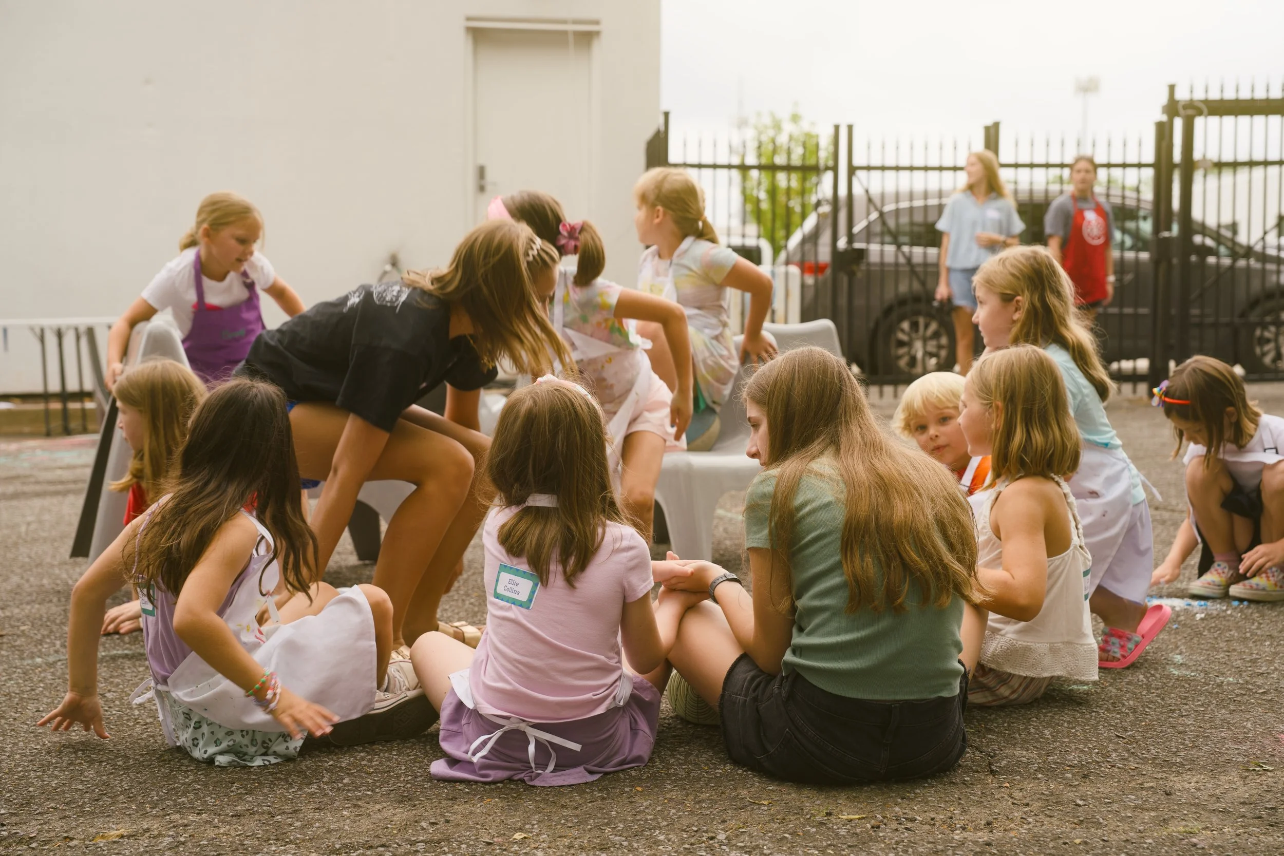 Group of young girls and women gathered outdoors, sitting on the ground and around a table, engaging in an activity, with a black fence and parked cars in the background.