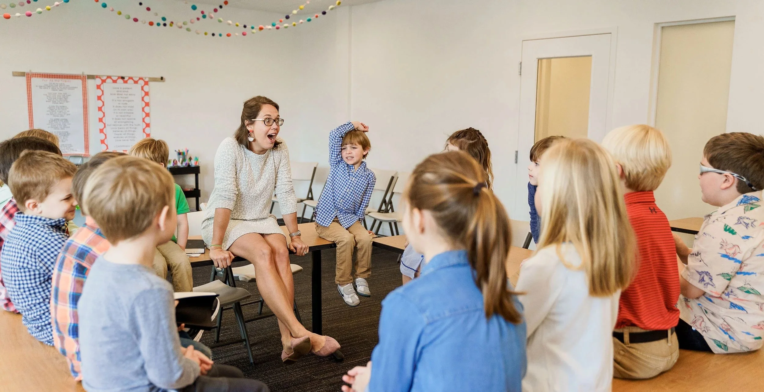 A teacher sits on a desk surrounded by children in a classroom, with one boy raising his hand, engaging in a lively discussion.