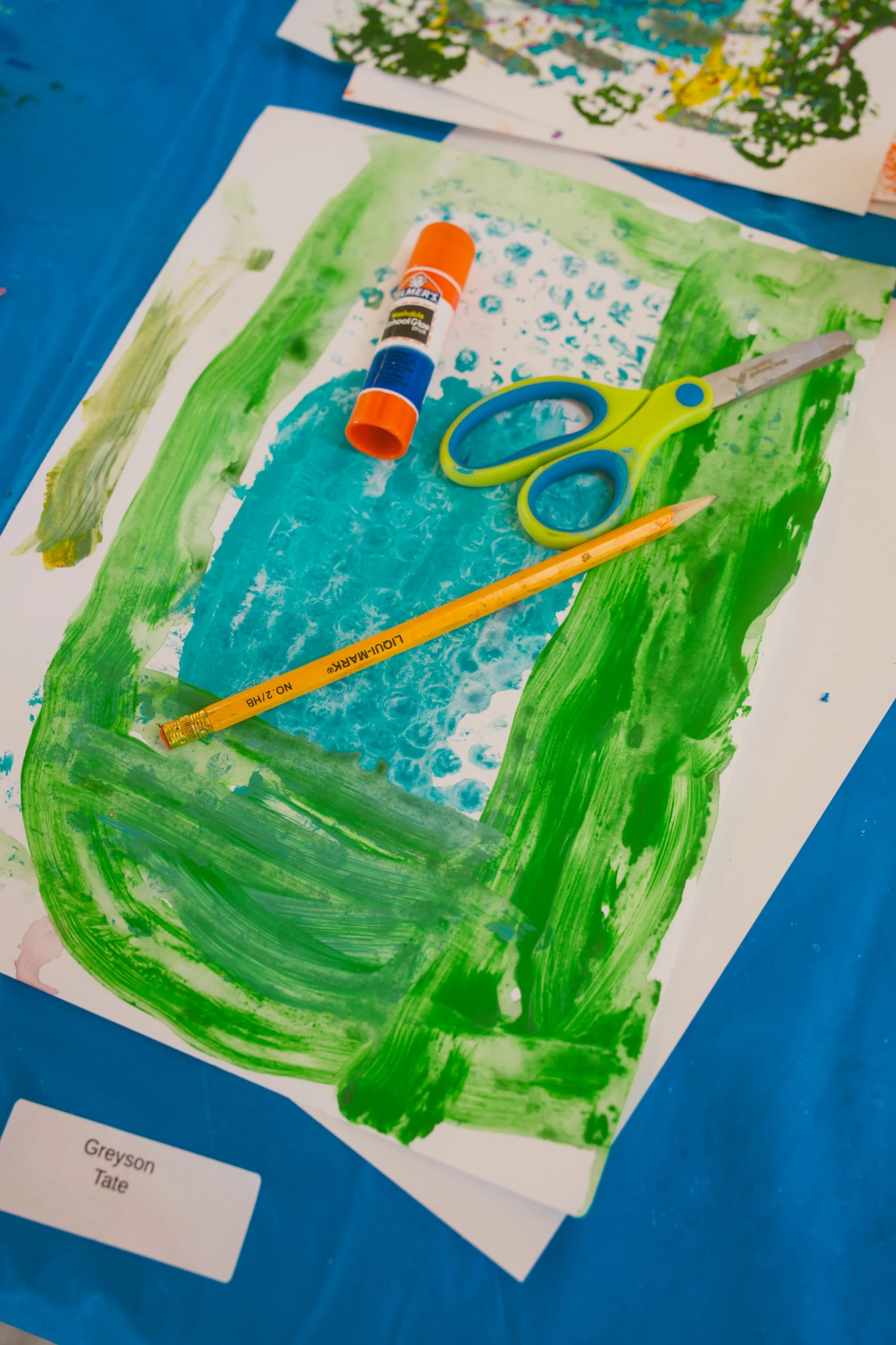 Colorful artwork on paper with green and blue paint, a pair of scissors, a glue stick, and a yellow pencil on a blue surface, likely part of an art project.