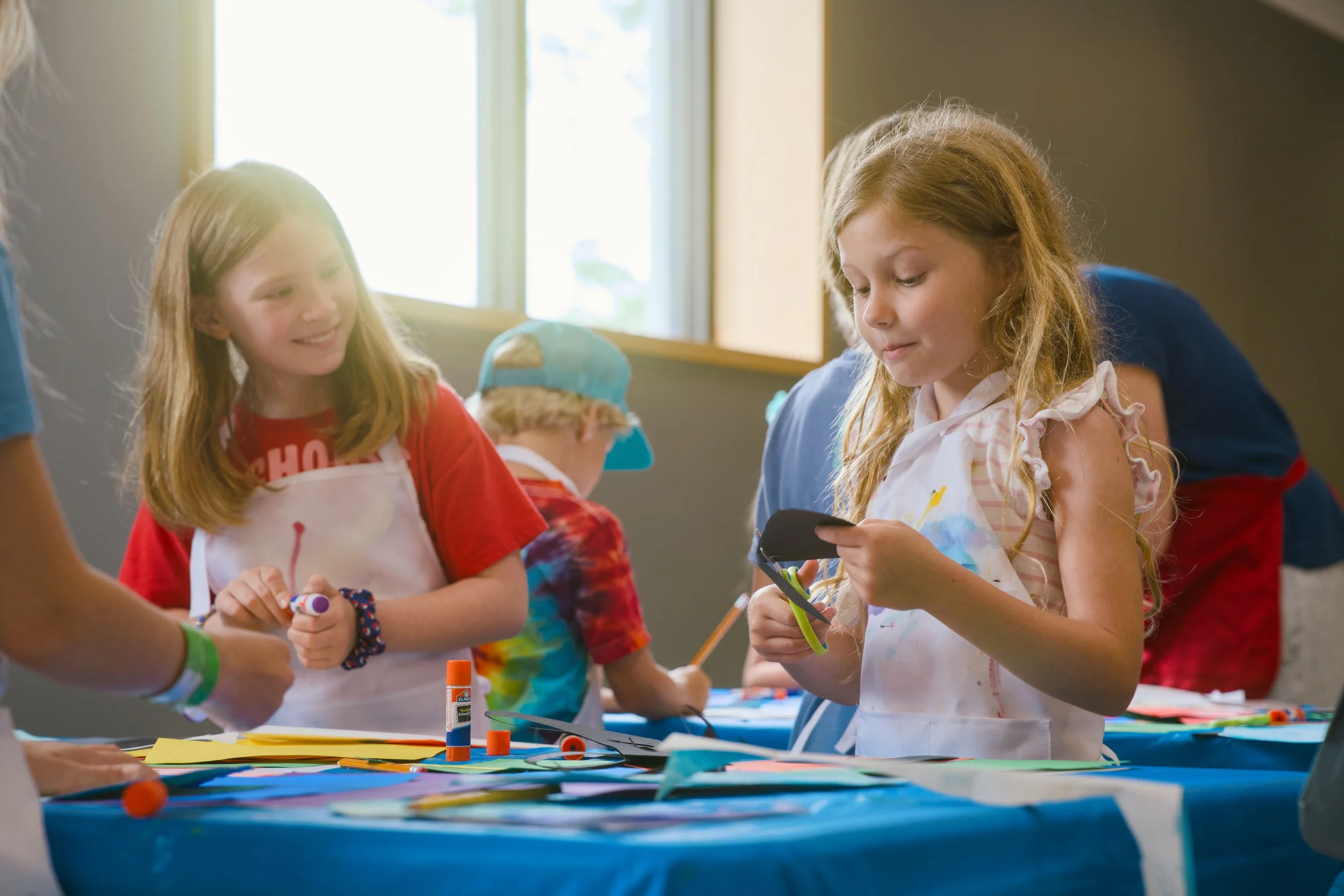 Group of children at an art and craft activity table, with art supplies and paper, in a brightly lit room.
