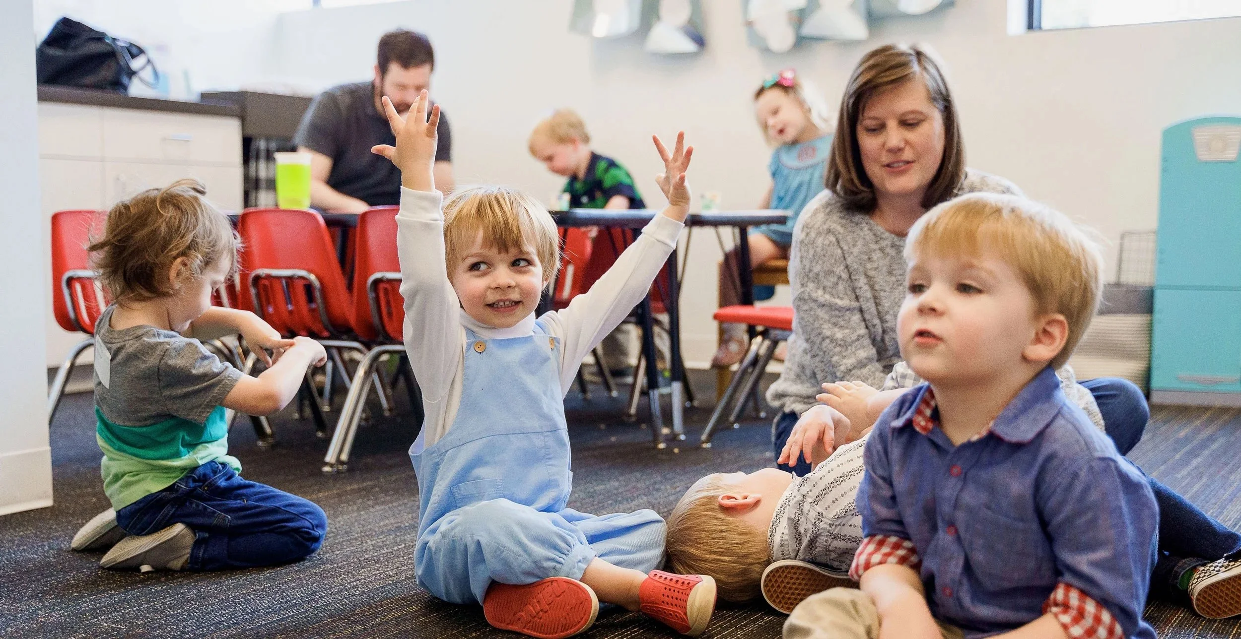 Children and adults participating in an indoor activity, with one child raising her hands while seated on the floor.