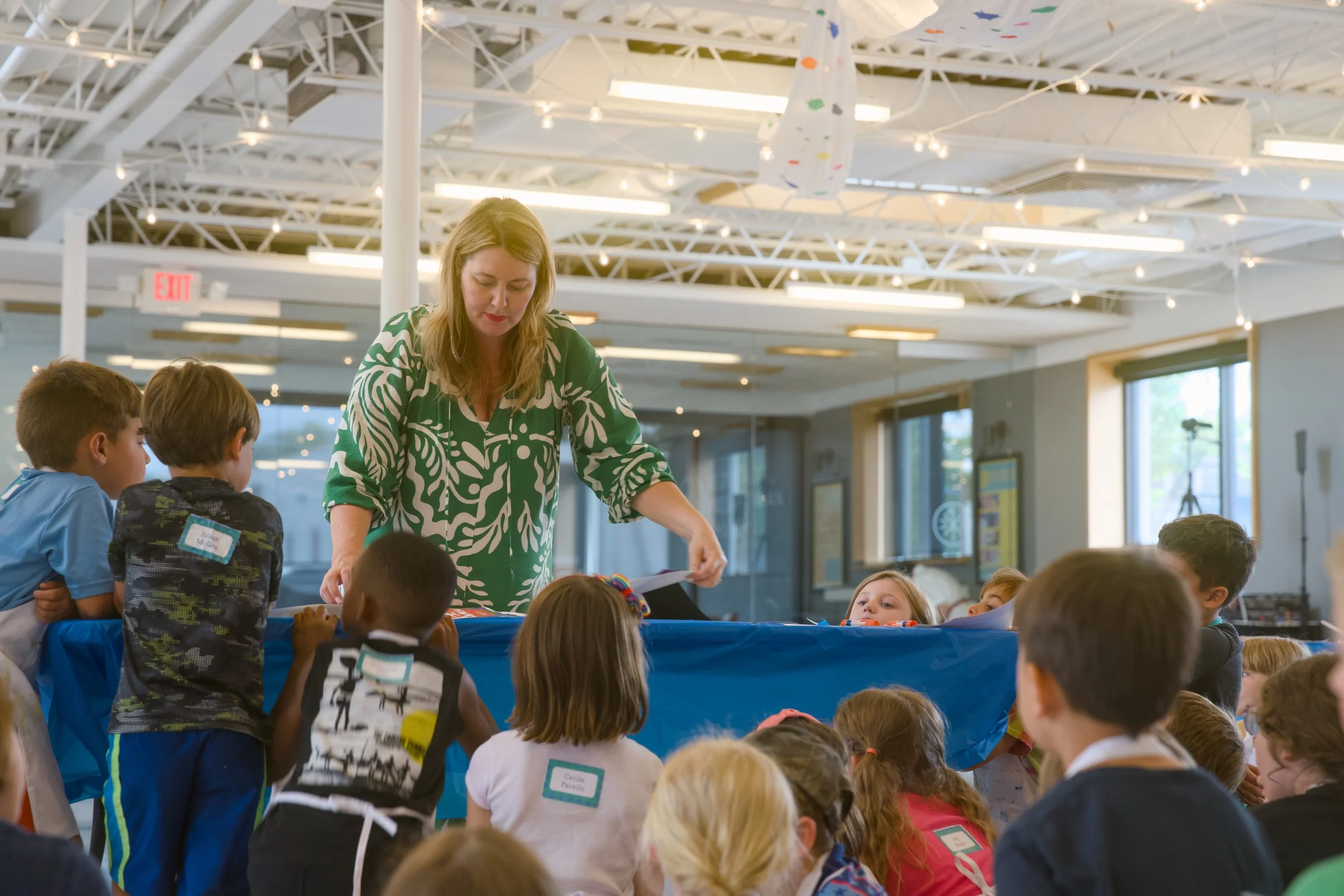 A woman in a green patterned blouse interacts with a group of children, who are seated around a table in a well-lit indoor space with high ceilings and string lights.