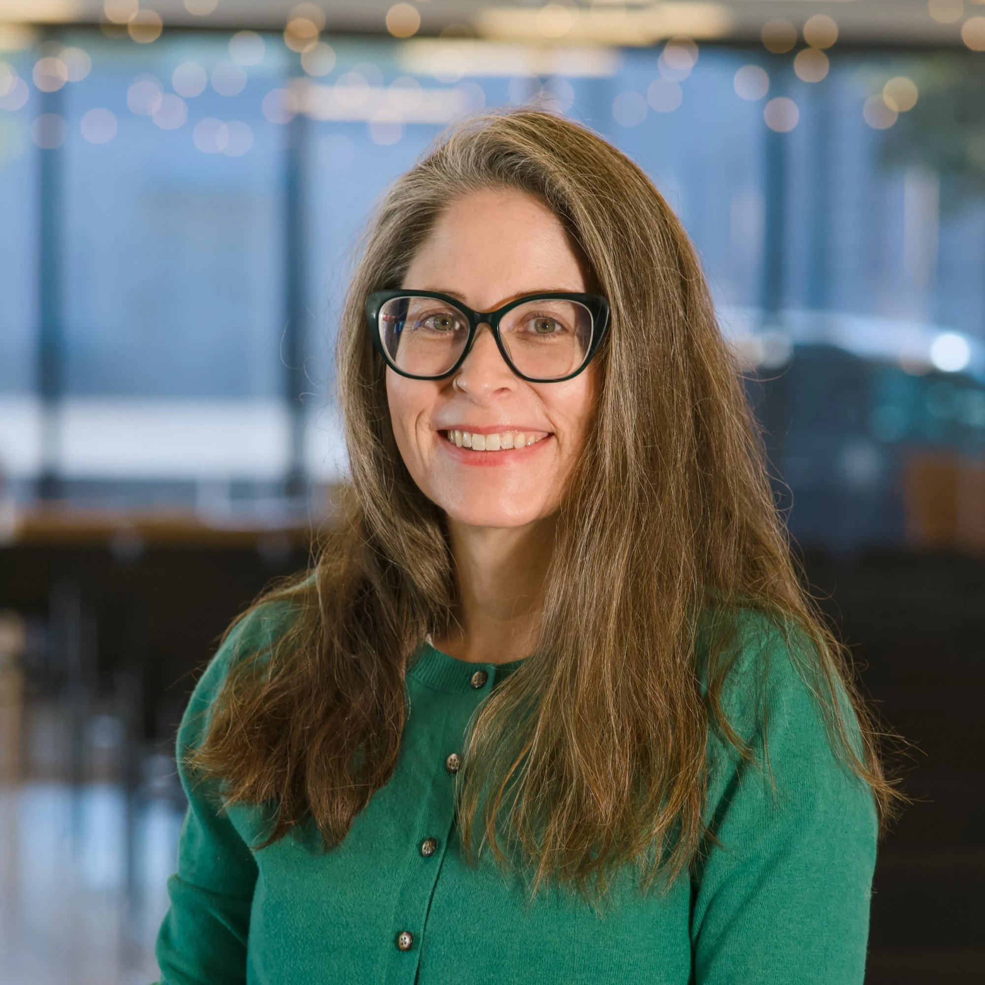 Close-up of woman with long brown hair, black glasses, wearing a green sweater, smiling indoors with blurred background.