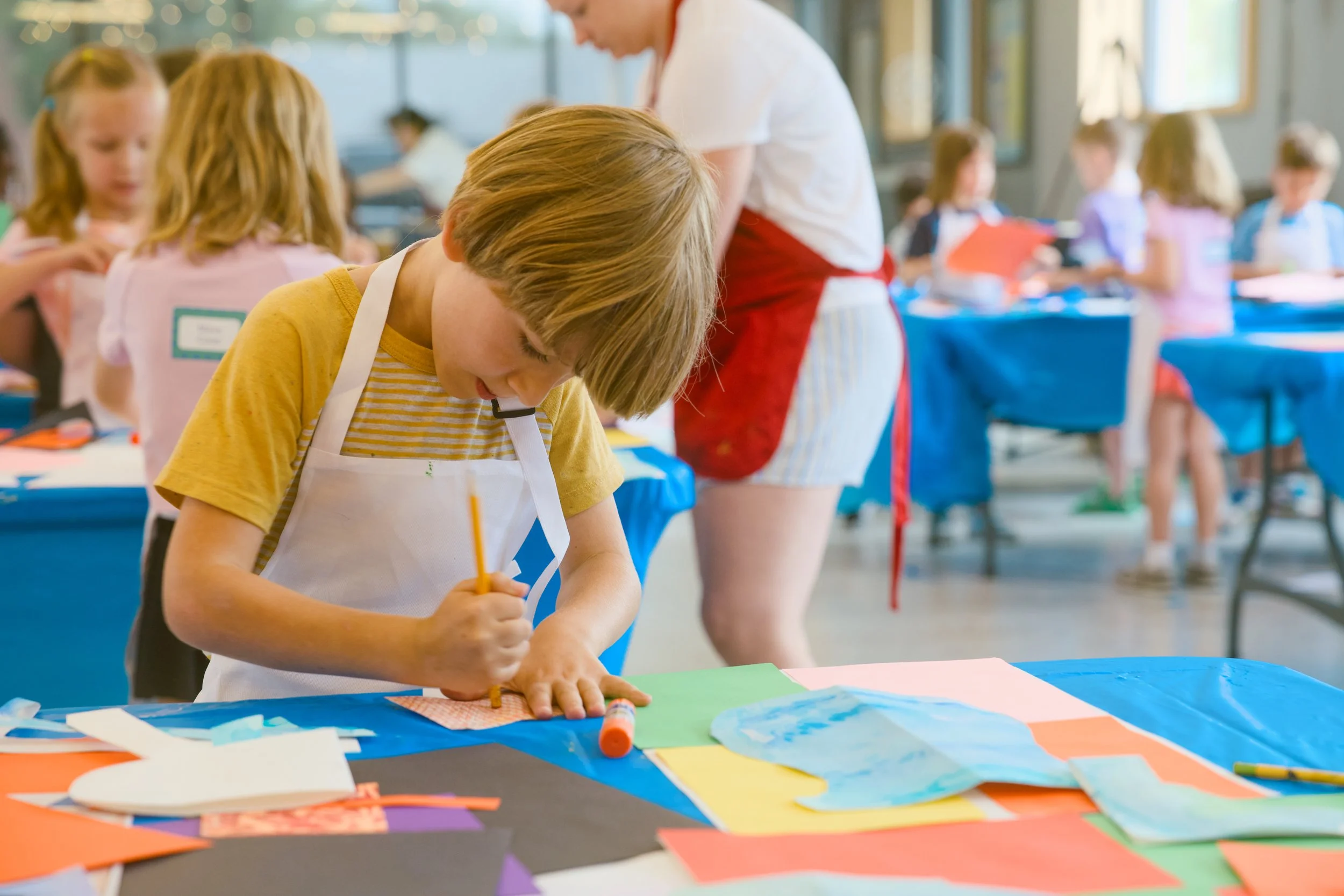 Child with light brown hair and a yellow shirt working on an arts and crafts project at a table filled with colorful paper and craft supplies in a bright room with other children and an adult in the background.