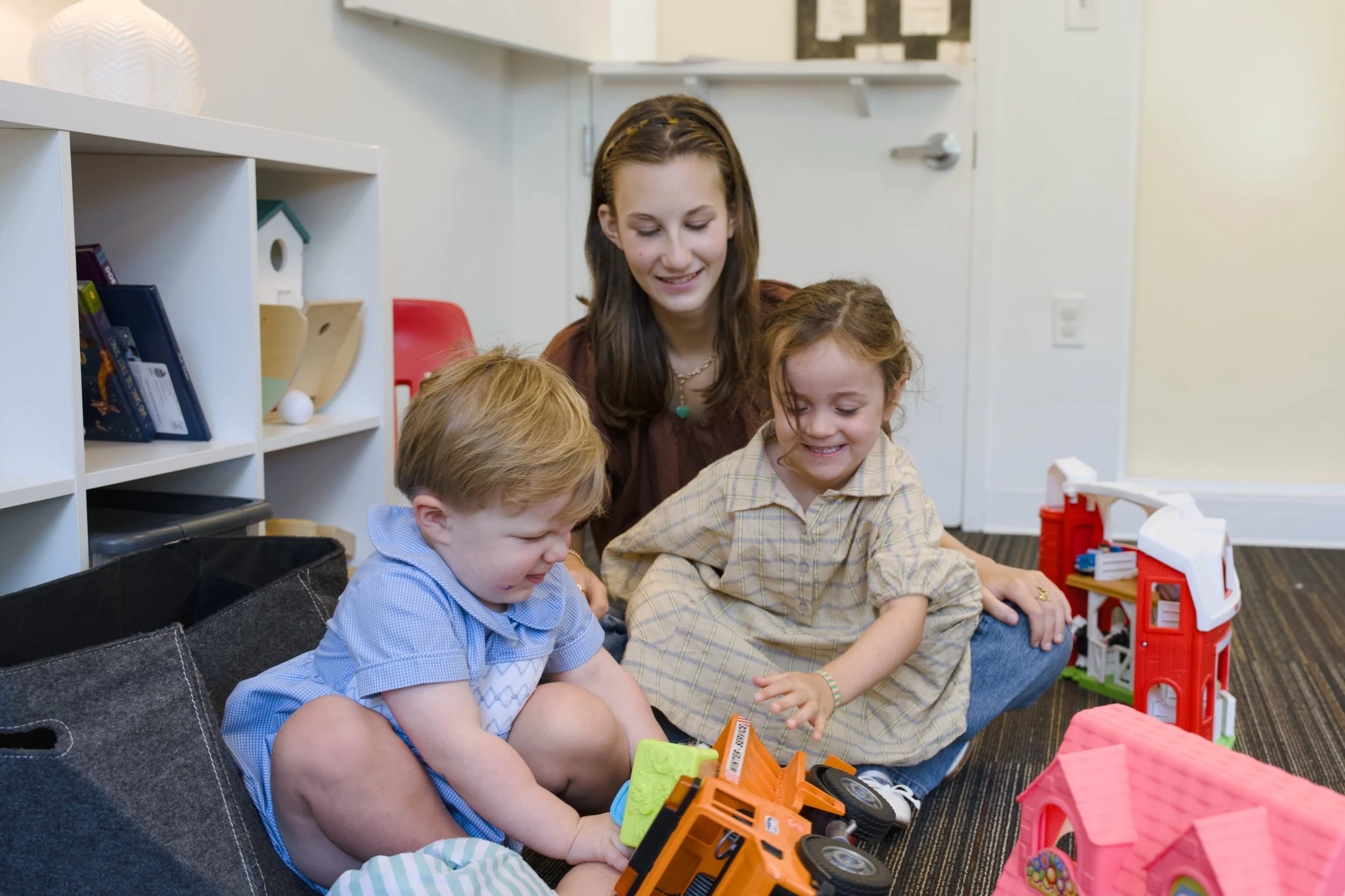 A woman and three young children are sitting on the floor playing with toy trucks and building blocks in a room with a white bookshelf, toys, and a door in the background.