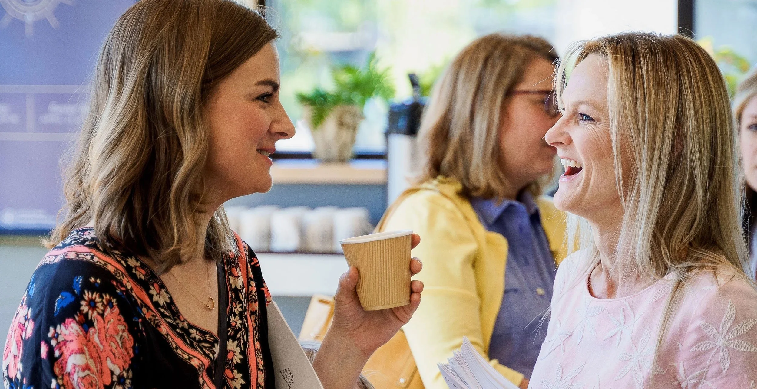Two women are smiling and engaging in conversation, one holding a paper cup.
