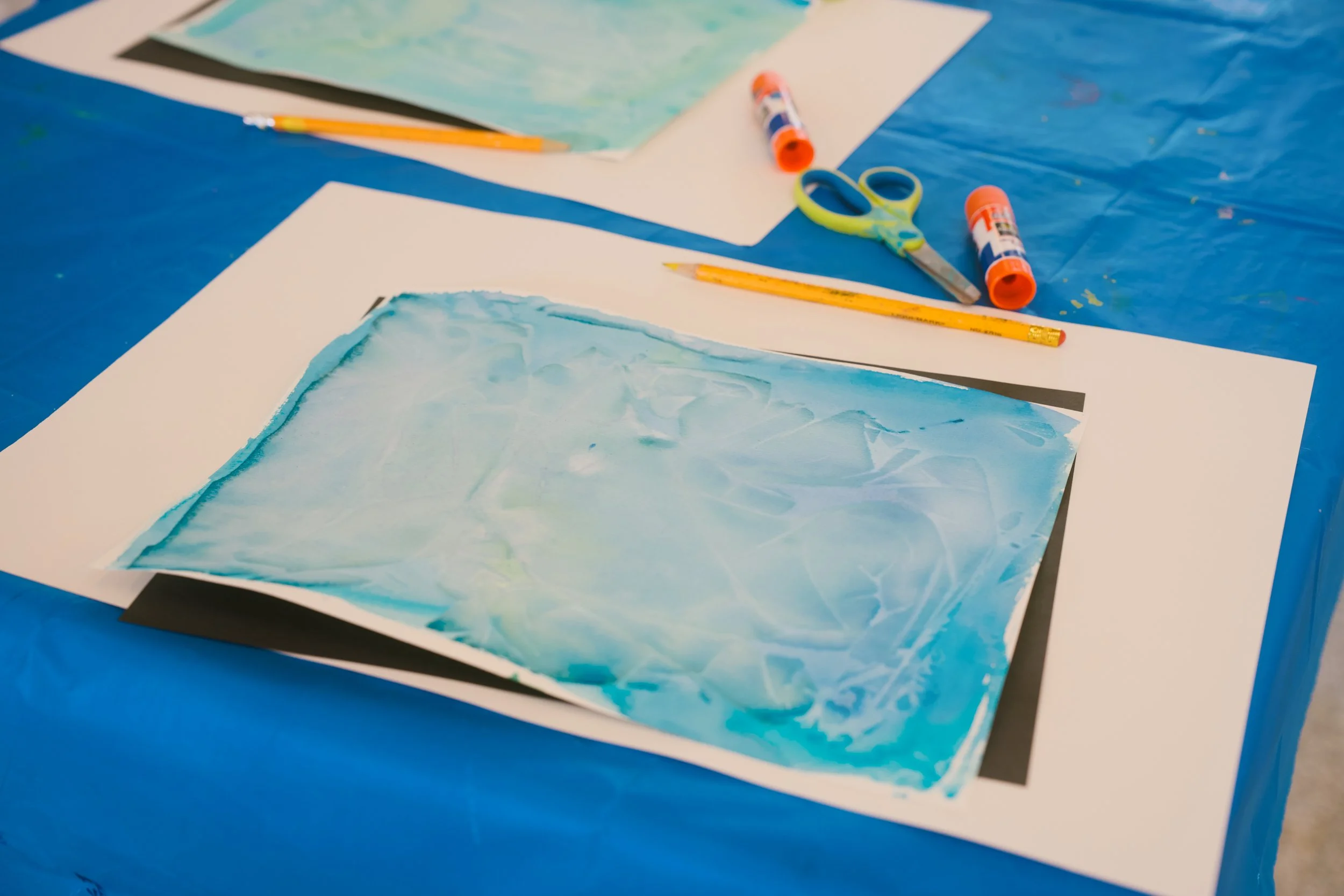 Table covered with blue tablecloth holding watercolor paintings, yellow pens, scissors, glue sticks, and paper.