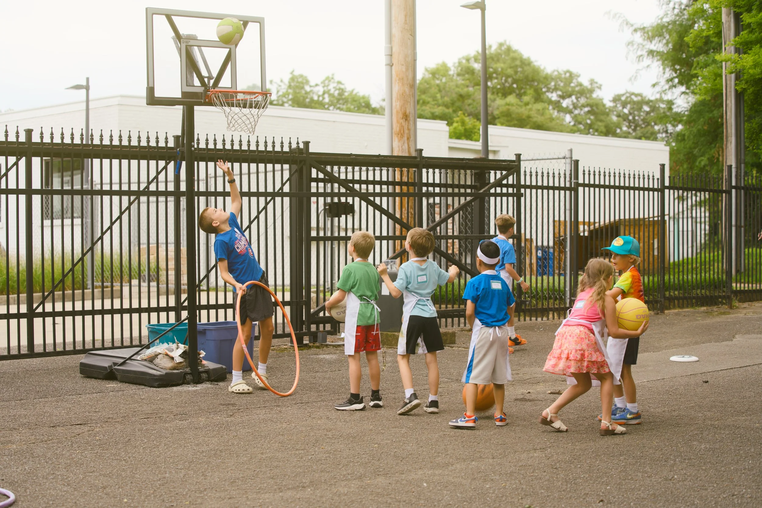 Children at an outdoor basketball court playing and talking, with some holding sports equipment and others observing. A basketball is seen going into the hoop, and there are various sports balls and bags on the ground.