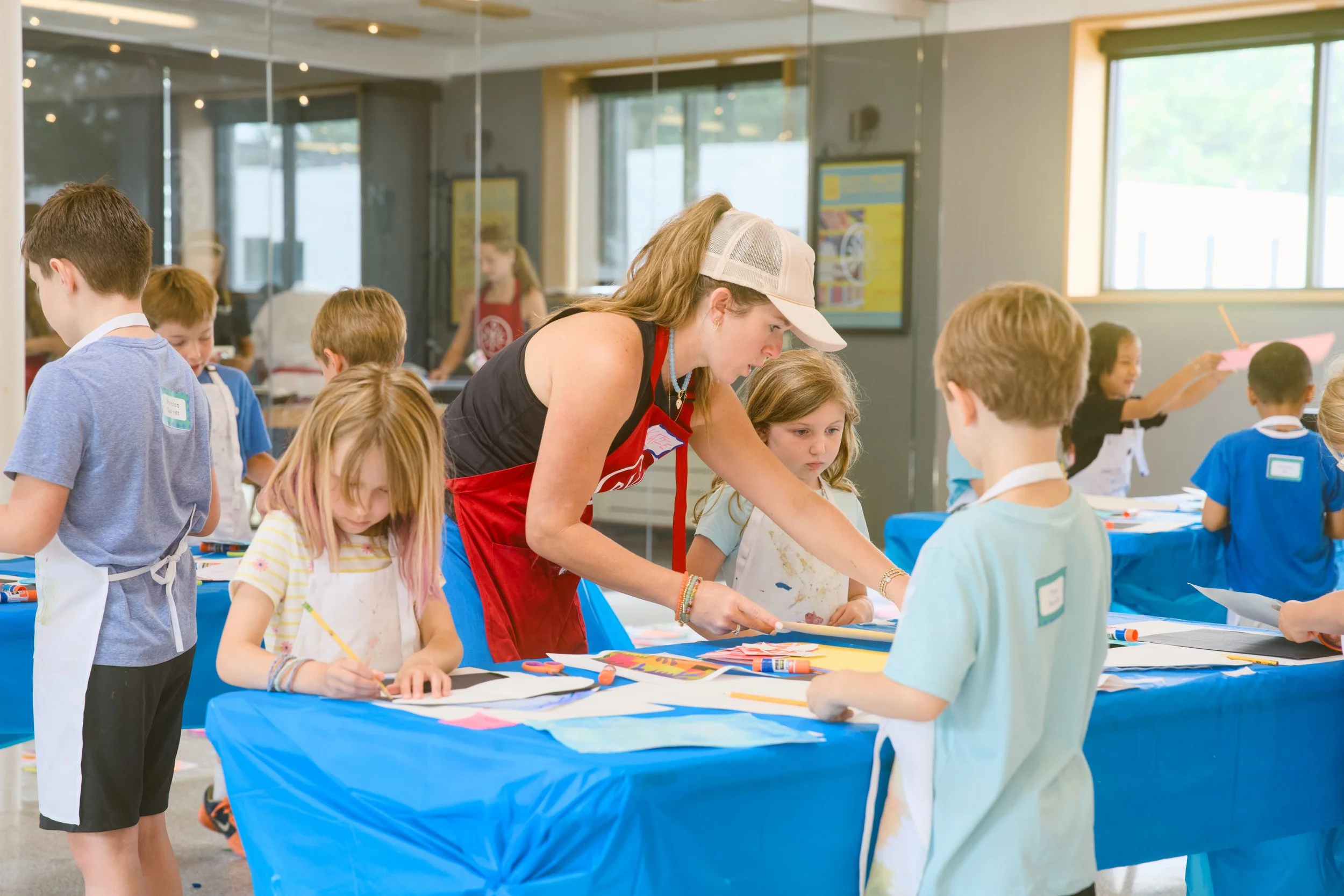 Children and an adult participating in an arts and crafts activity at a table with a blue tablecloth in a brightly lit room with large windows.