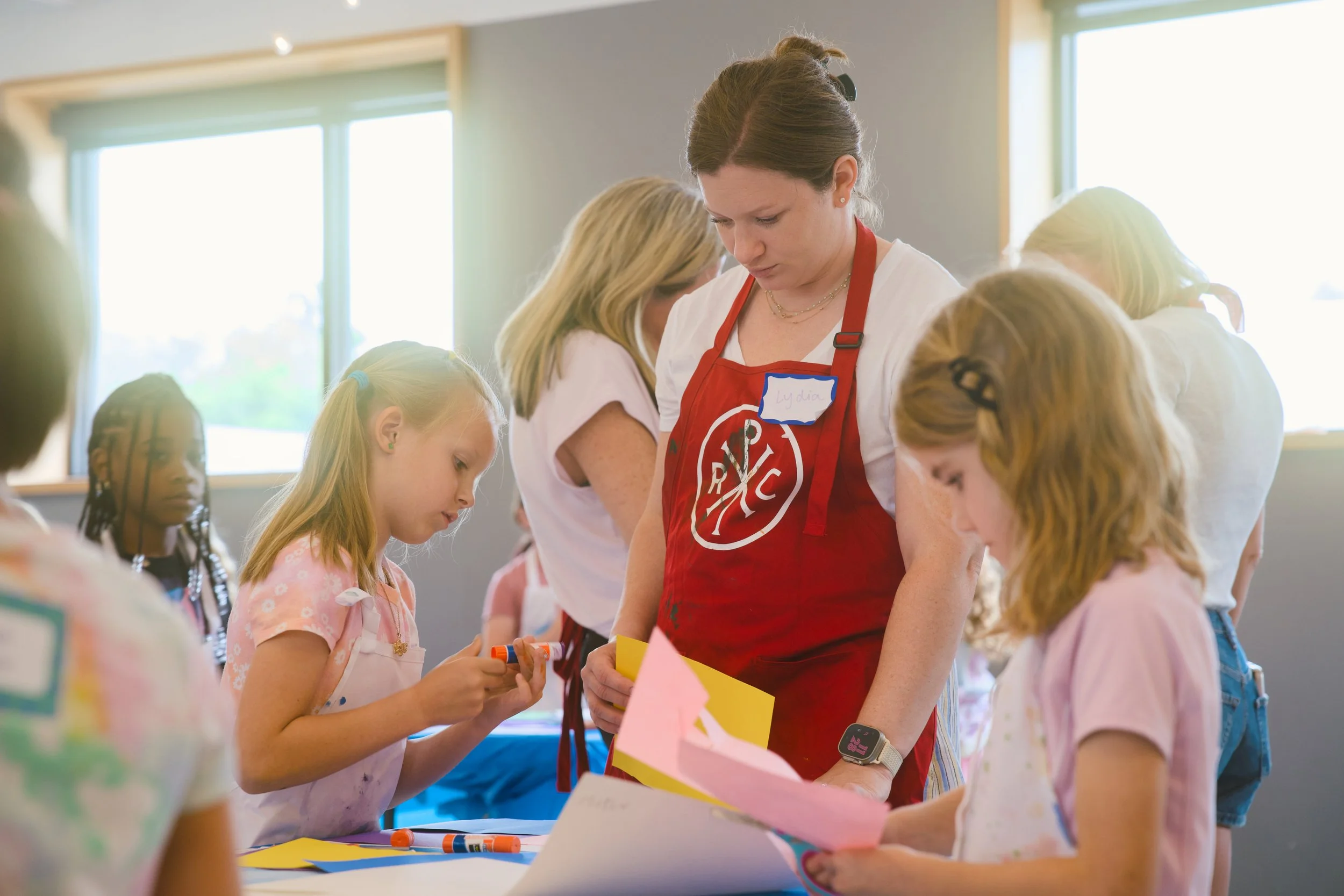 A group of children and a woman with a red apron working on arts and crafts at a table, with large windows in the background.