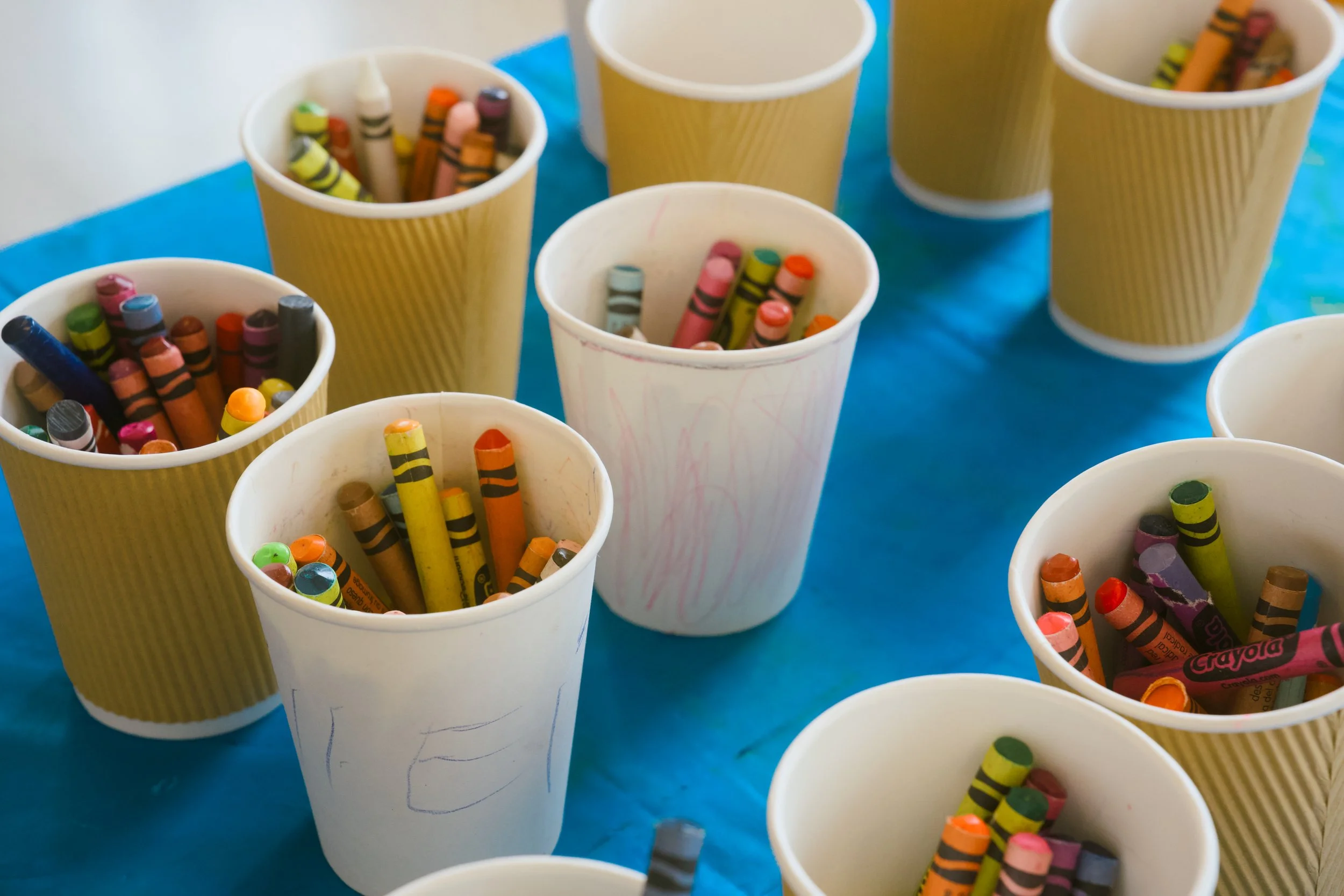 Several paper cups filled with colorful crayons on a blue table, some cups are decorated with children's drawings.