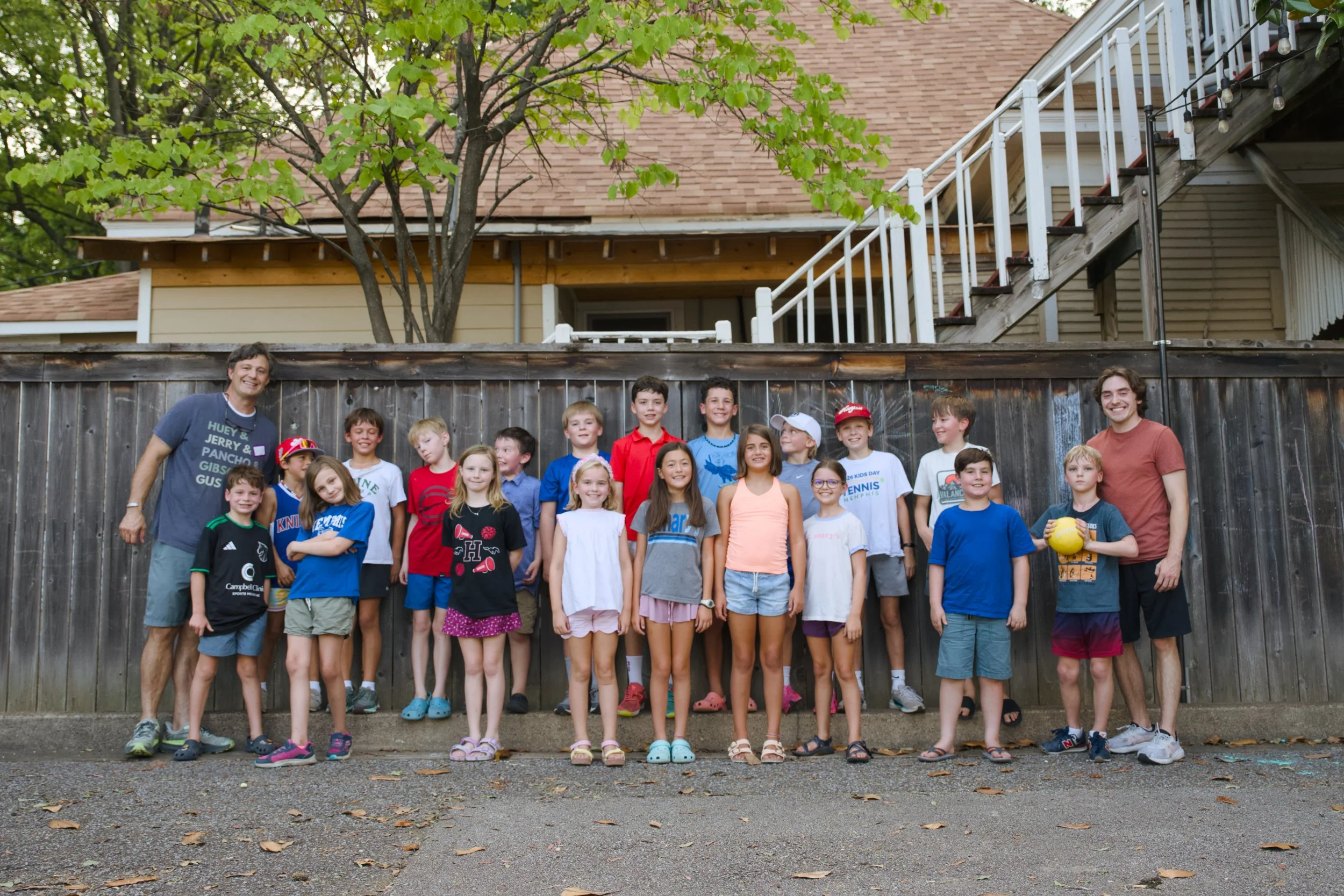 A group of children and two adult men posing for a photo outdoors in front of a wooden fence, with a house and a tree in the background. The children are smiling and many are wearing colorful t-shirts and shorts.
