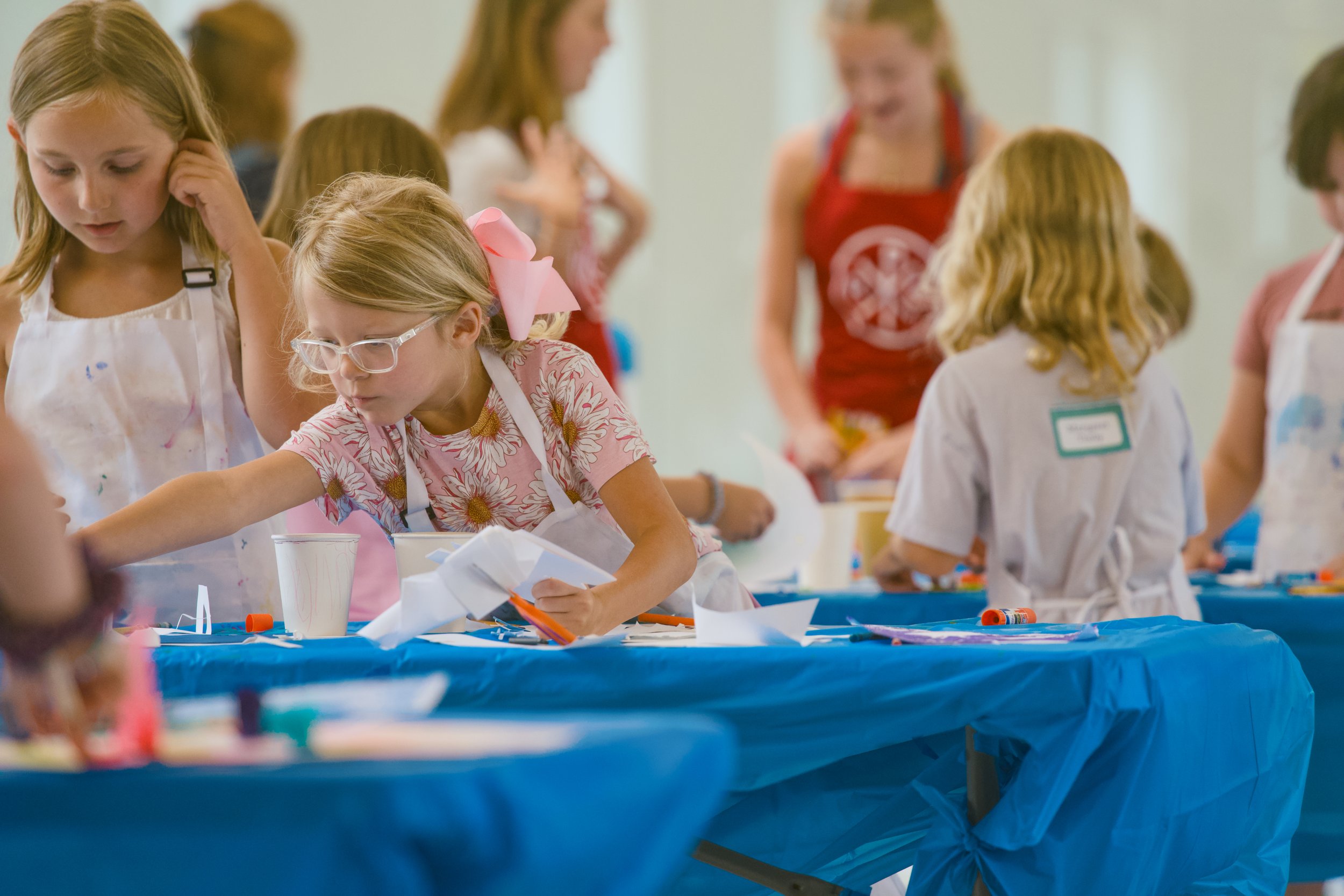 Kids and adults participating in arts and crafts at a table during an indoor event, wearing aprons.