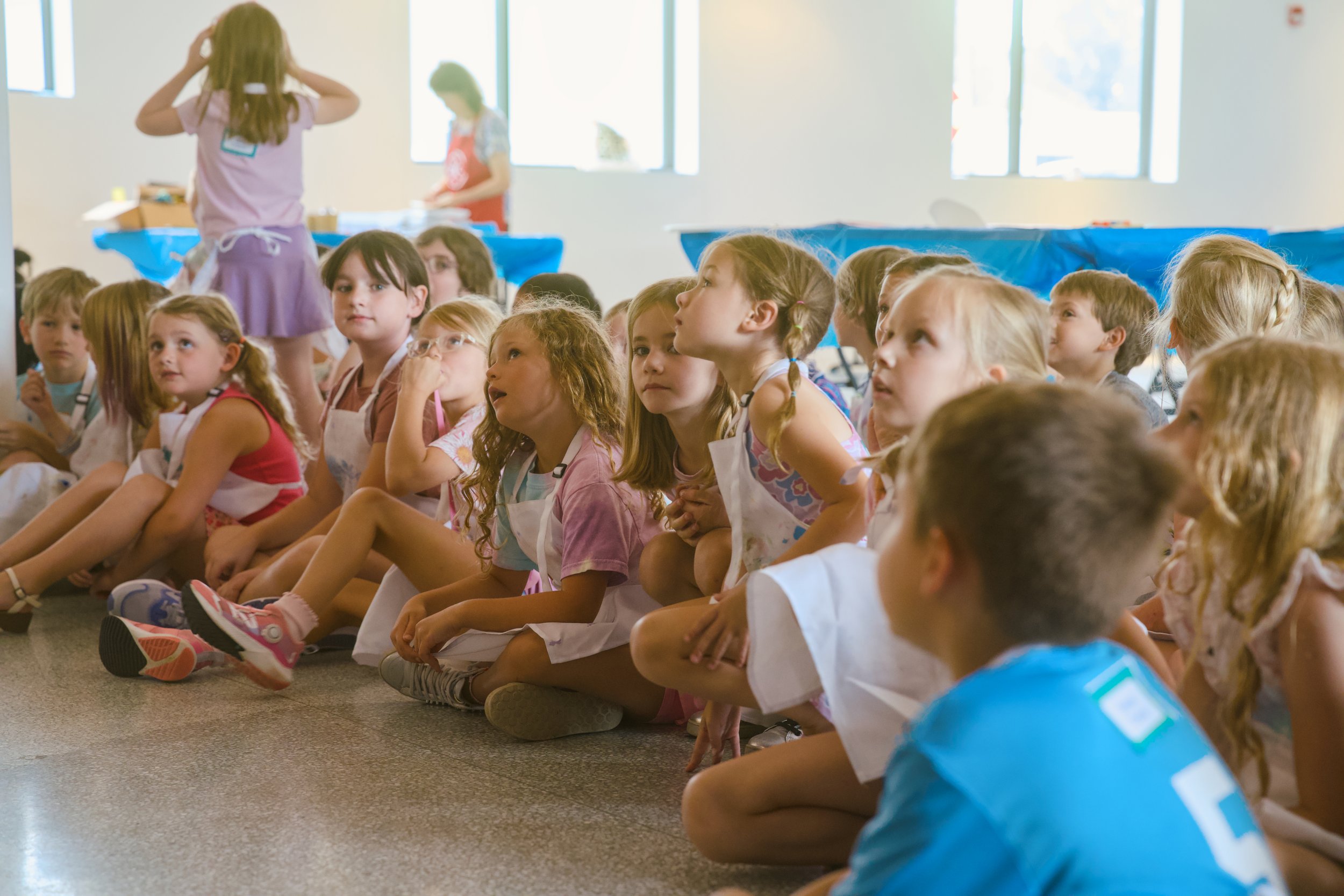 A group of children sitting on the floor, watching something with interest, as a woman in an apron prepares food behind them in a bright room with large windows.