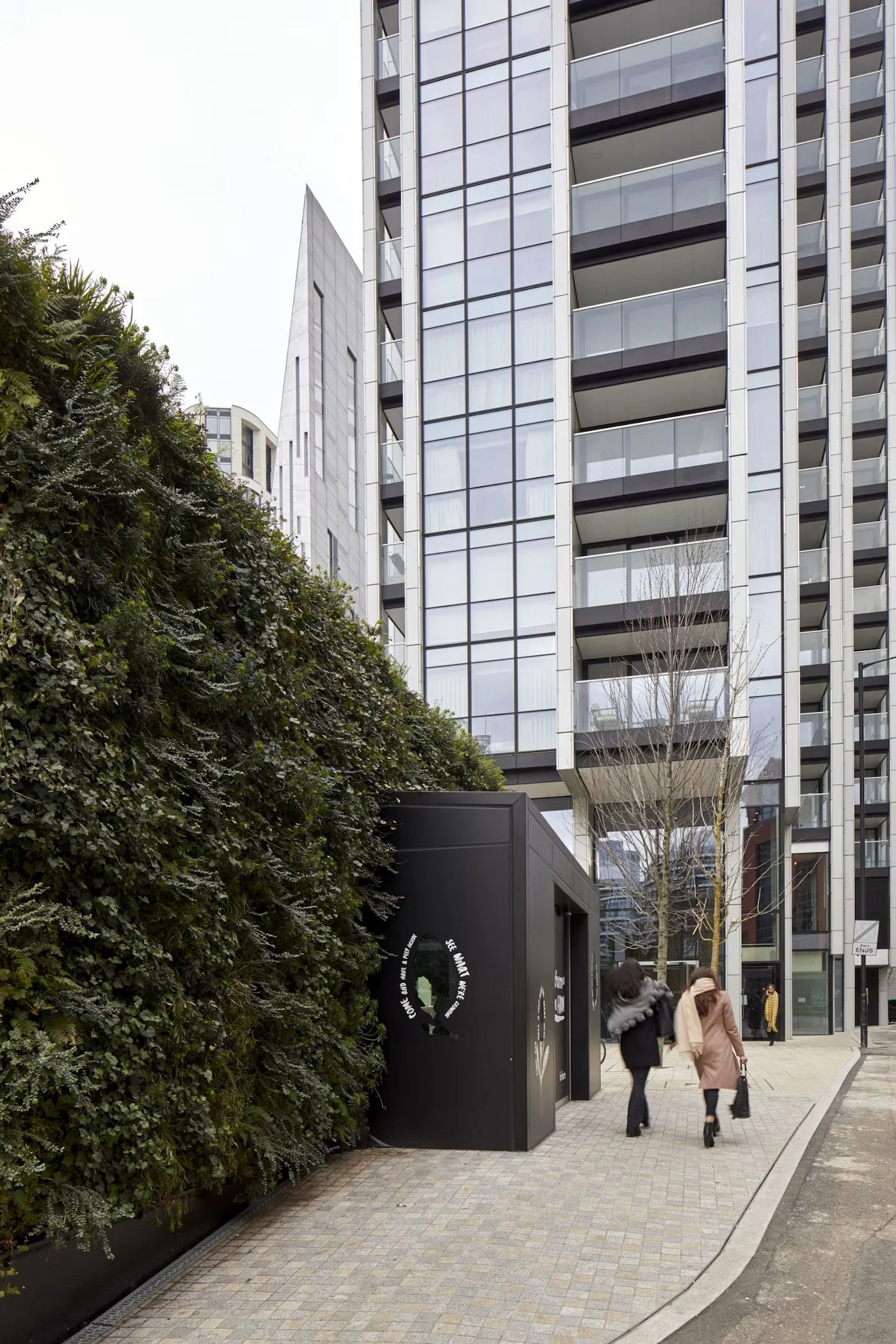 Tall modern glass and metal building with balconies, pedestrians walking on paved sidewalk, greenery on the left side, and a black structure with white text near the entrance.